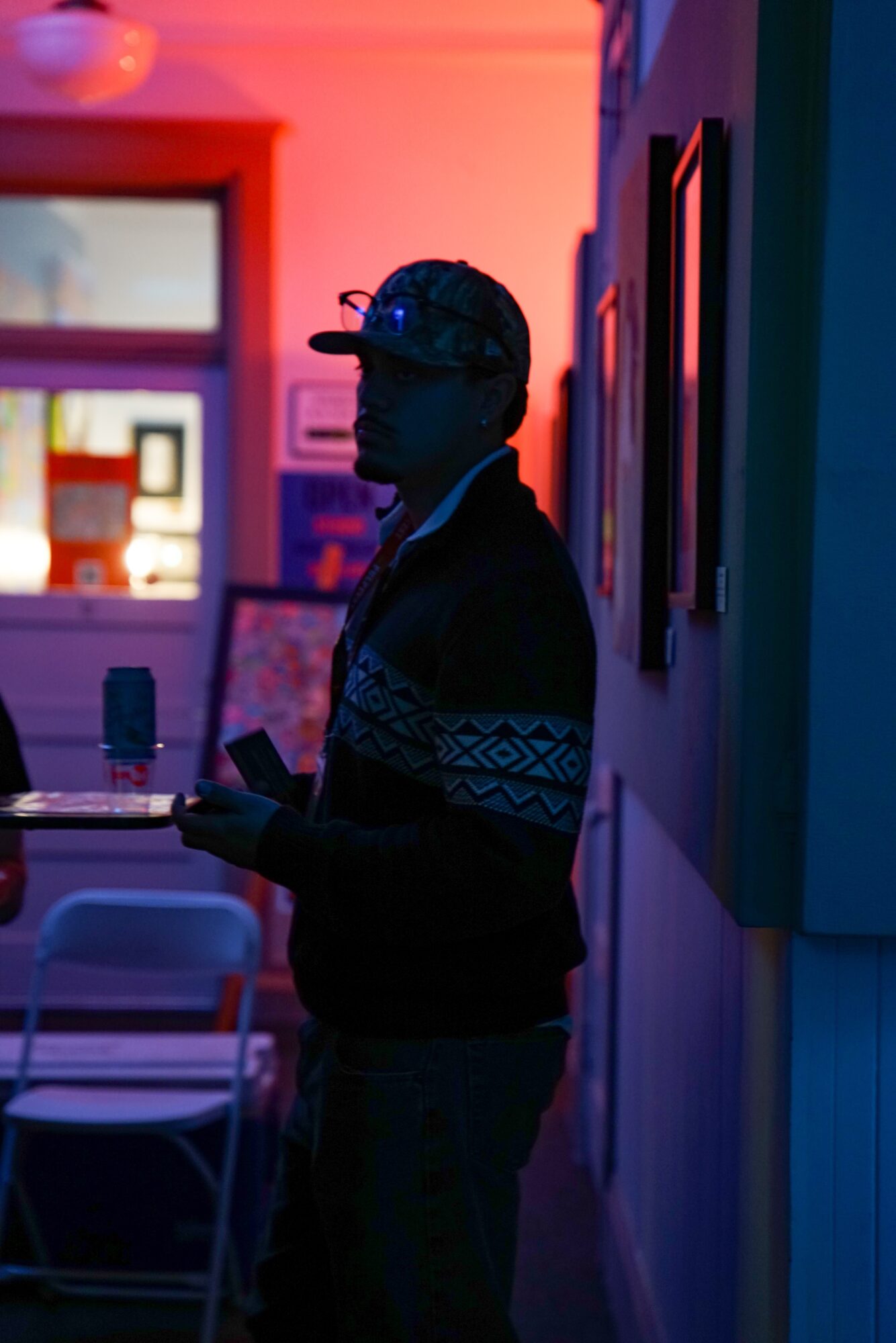 Silhouette of a person holding a phone in a dimly lit room with colorful lighting, standing near a wall with framed pictures.
