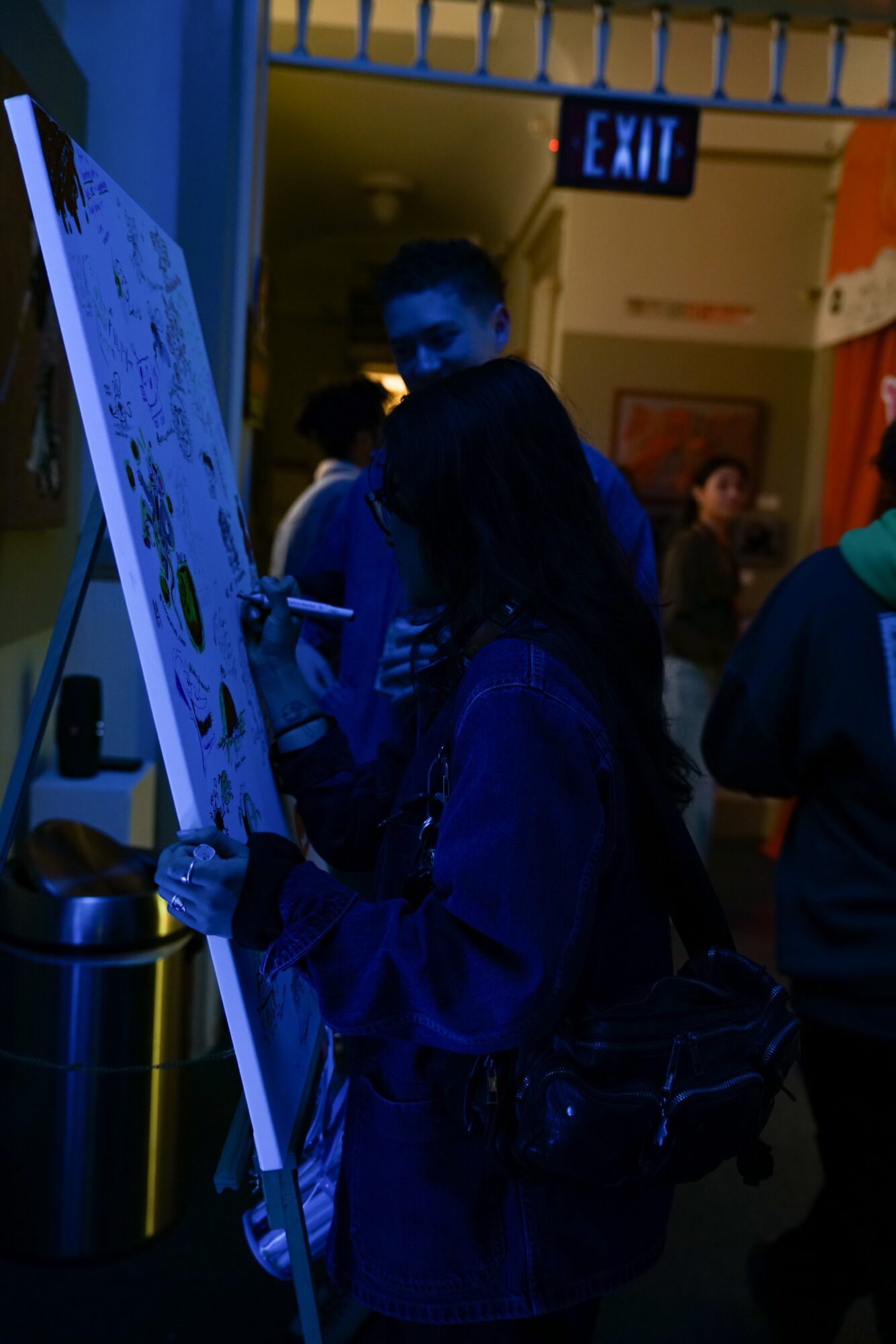 Person standing next to an informational board in a dimly lit indoor space with other people around.