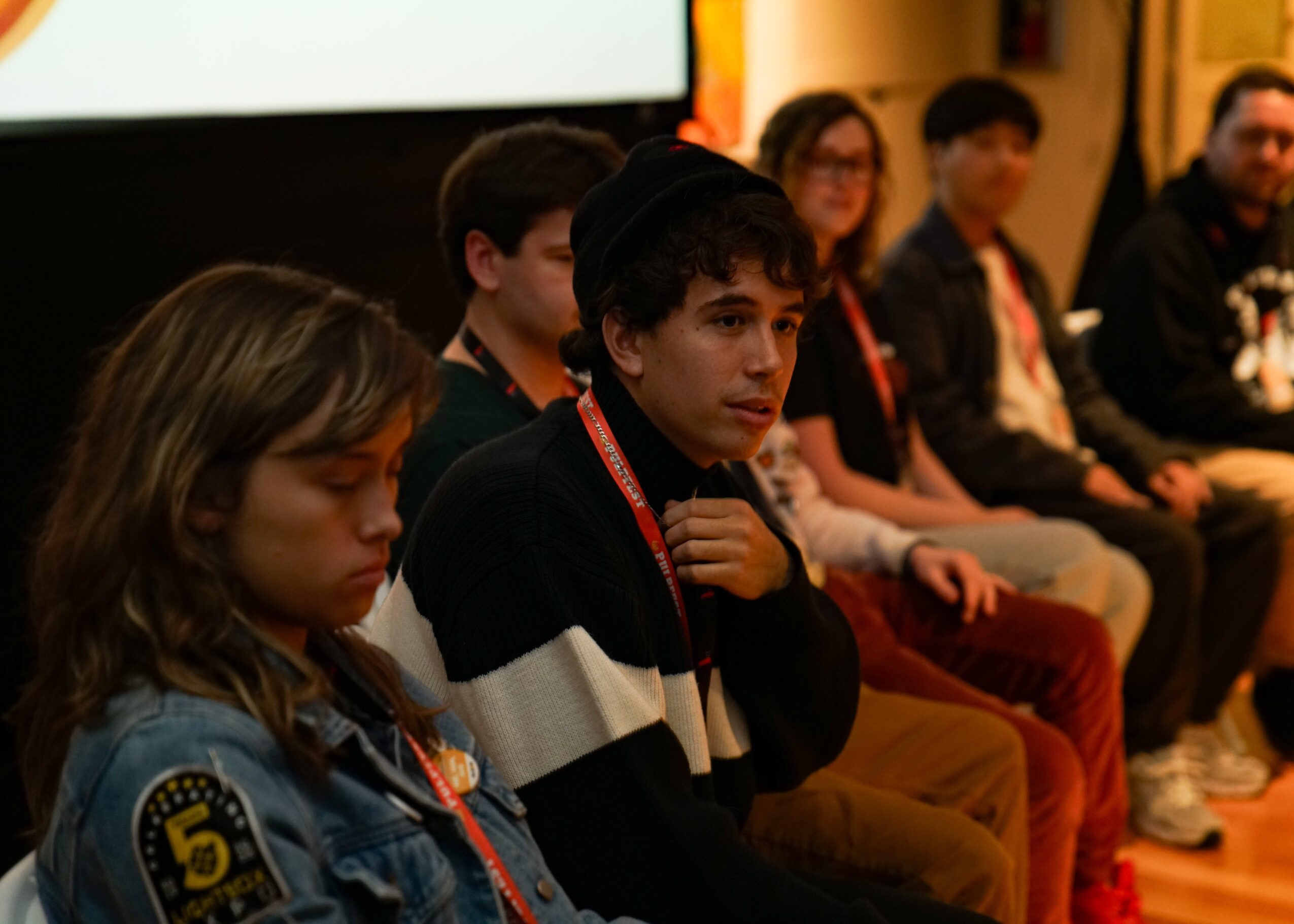 Group of young people sitting in a panel or discussion setting, some looking thoughtful or attentive.