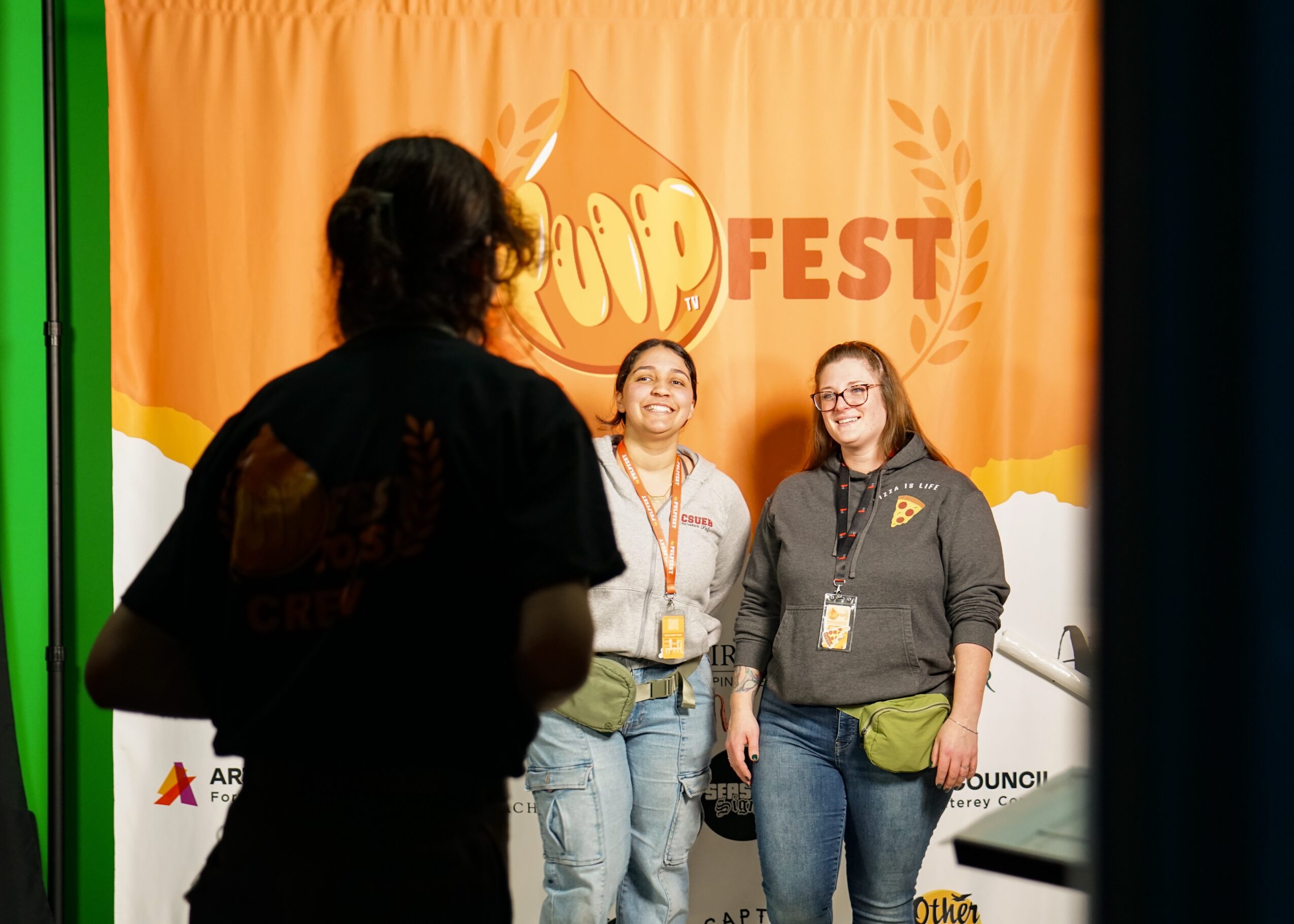 Two women standing on stage in front of a bright orange backdrop with the word 'POP FEST' and a popcorn graphic, smiling.