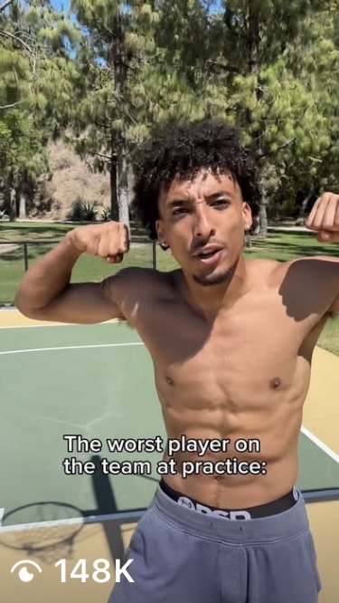 Young shirtless man flexing arms outdoors on a basketball court, trees in background, with caption about being the worst player.