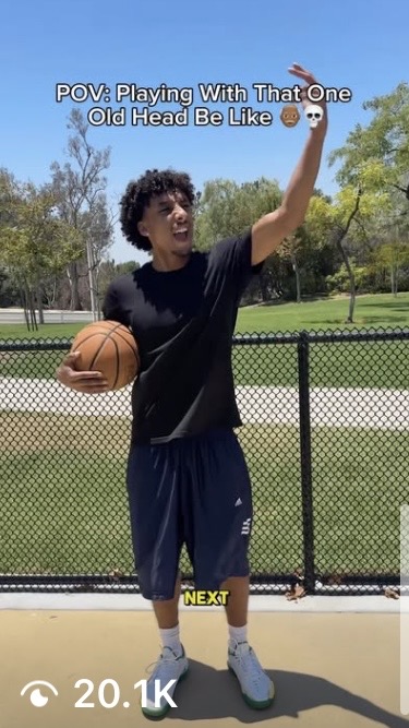 Young person holding basketball, pointing upward, outdoors on a sunny day, with trees and a fence in background.