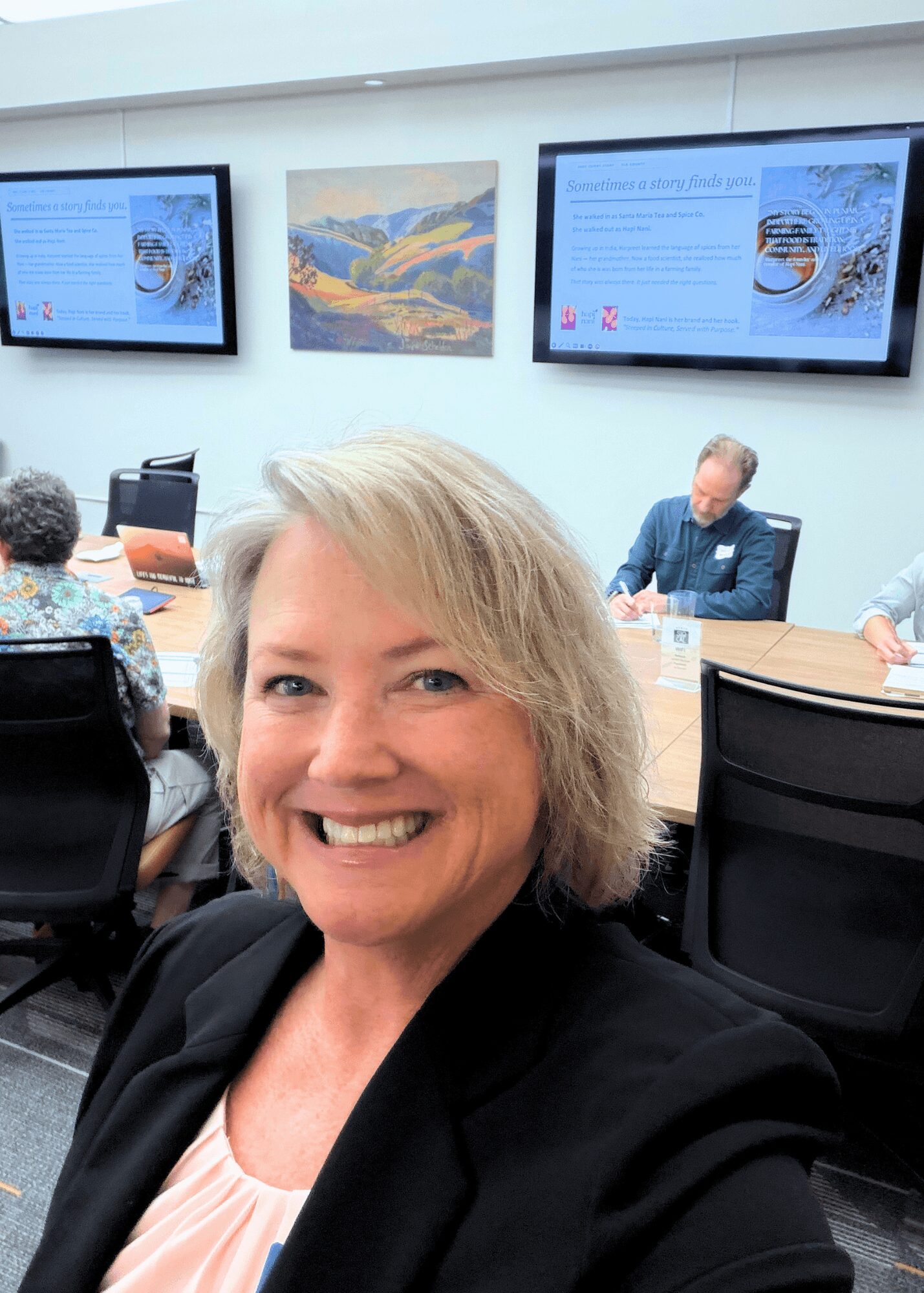 Smiling woman with blonde hair taking a selfie in a conference room with screens and people in the background.