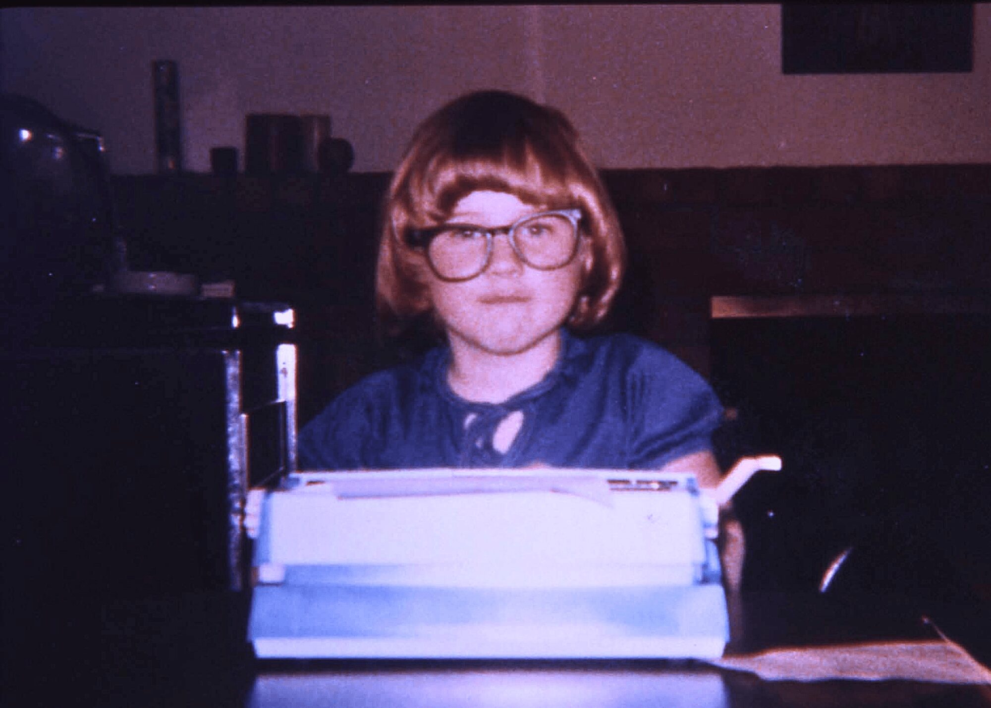 Child with glasses and short hair sitting at a desk with a typewriter, in a dimly lit room.