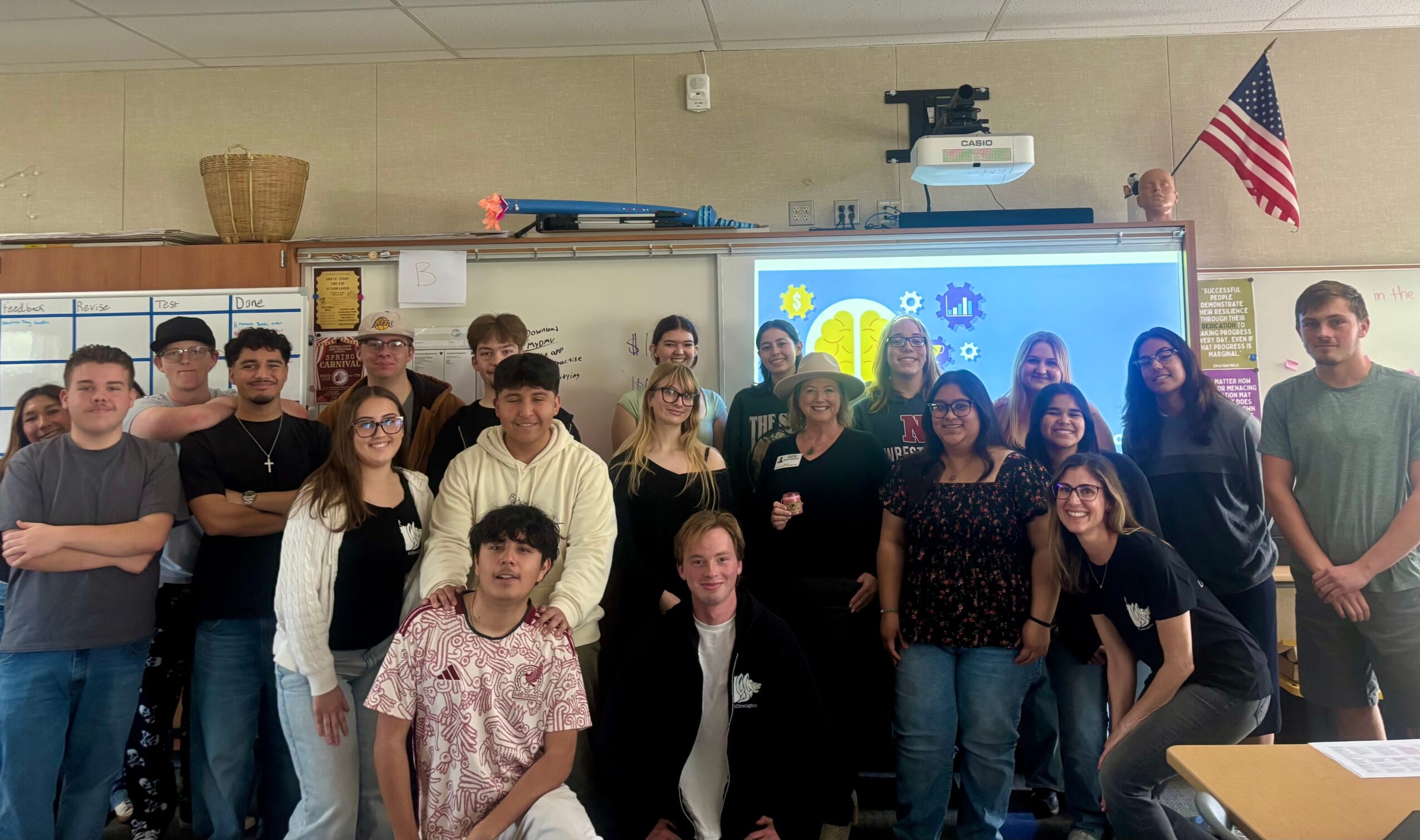 Group of students and teacher in classroom, some smiling, standing in front of a digital screen with a sun and cloud graphic.