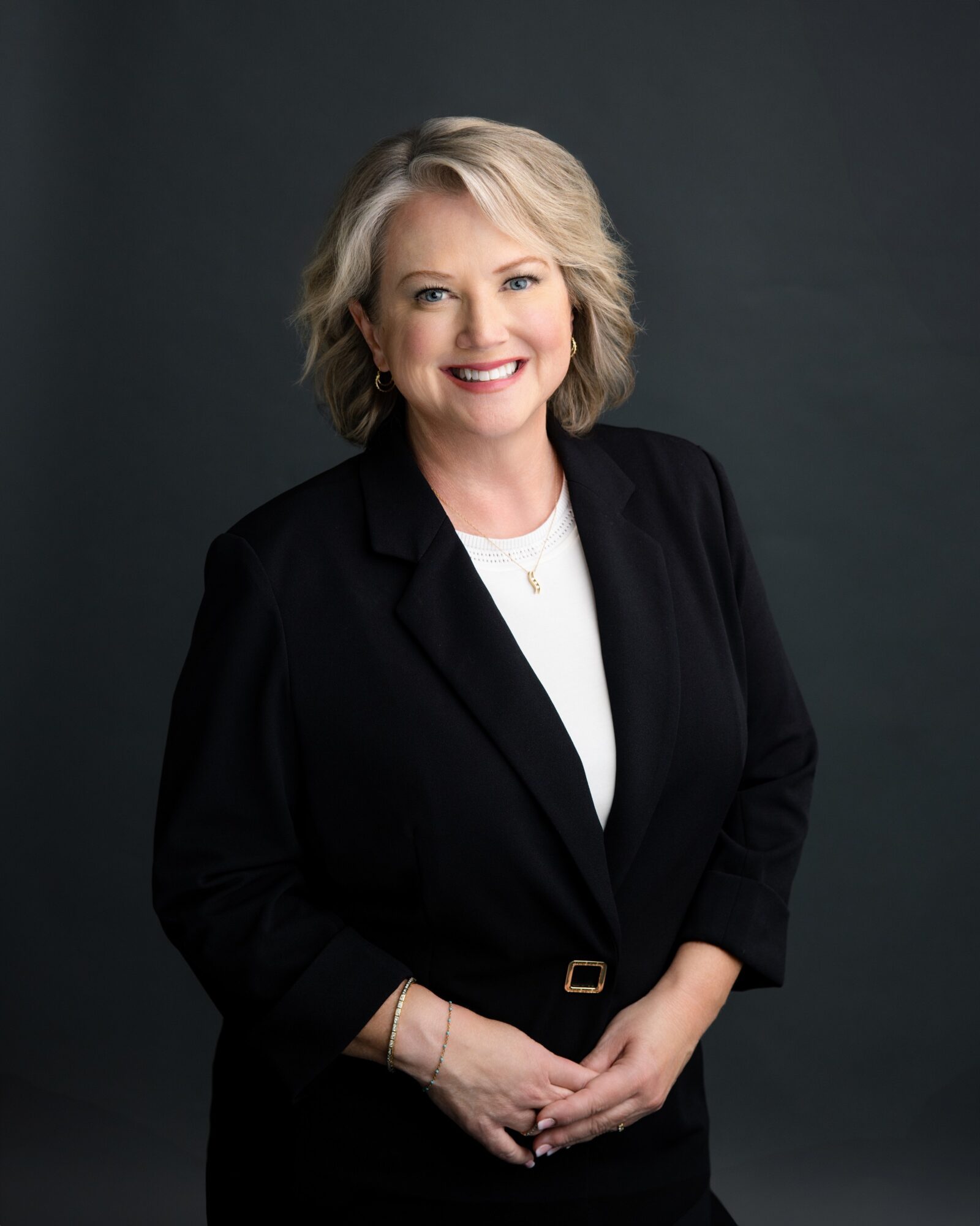 Smiling woman with blonde hair in a black blazer and white top, standing against a dark background.