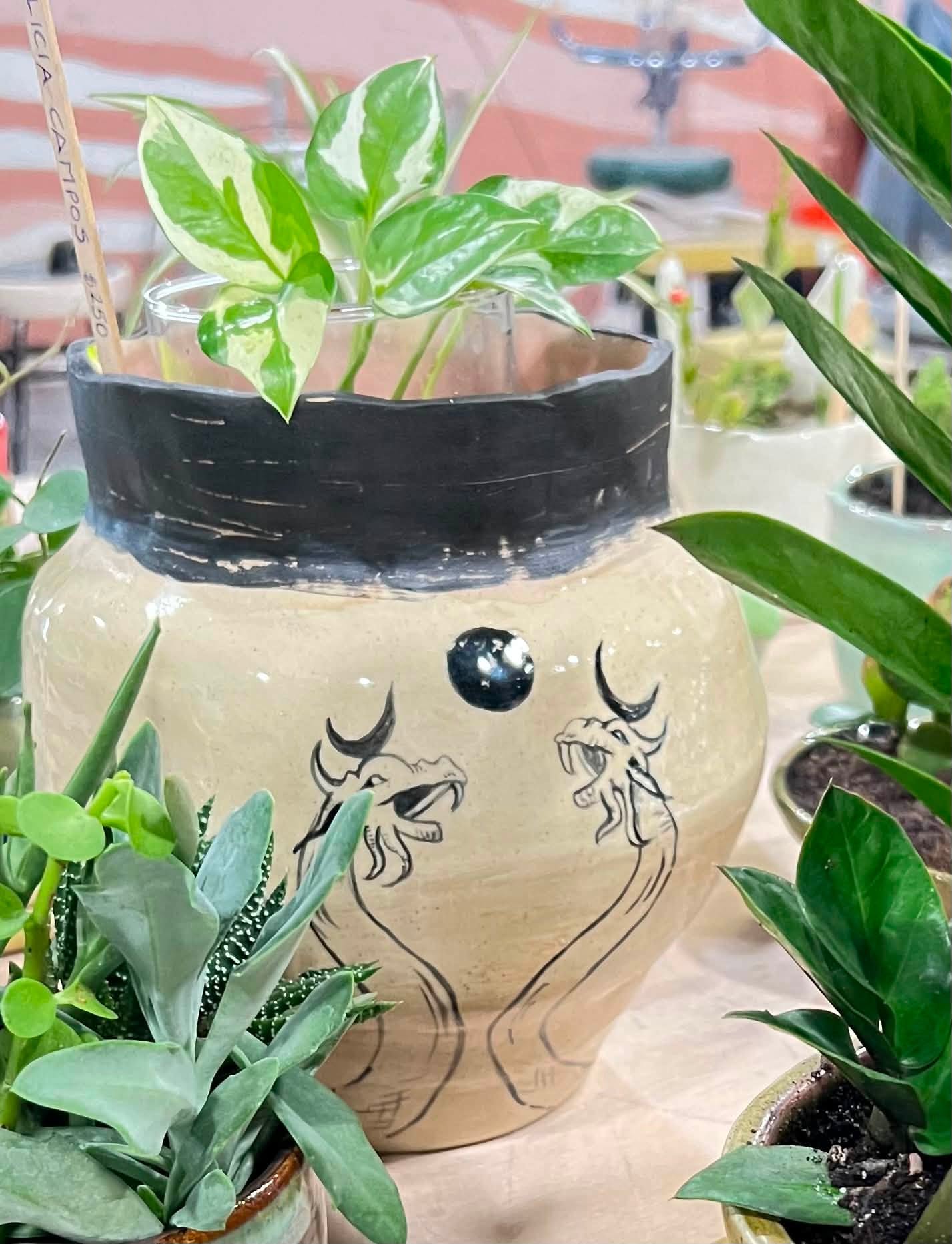 Decorative ceramic pot with painted face and plants inside, surrounded by other potted plants on a surface.