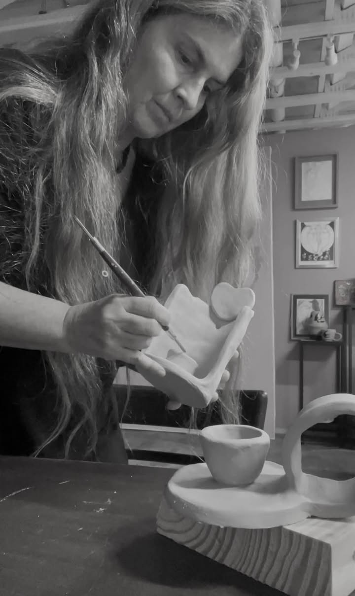 Woman with long hair painting on a ceramic object in a room with framed pictures on the wall.