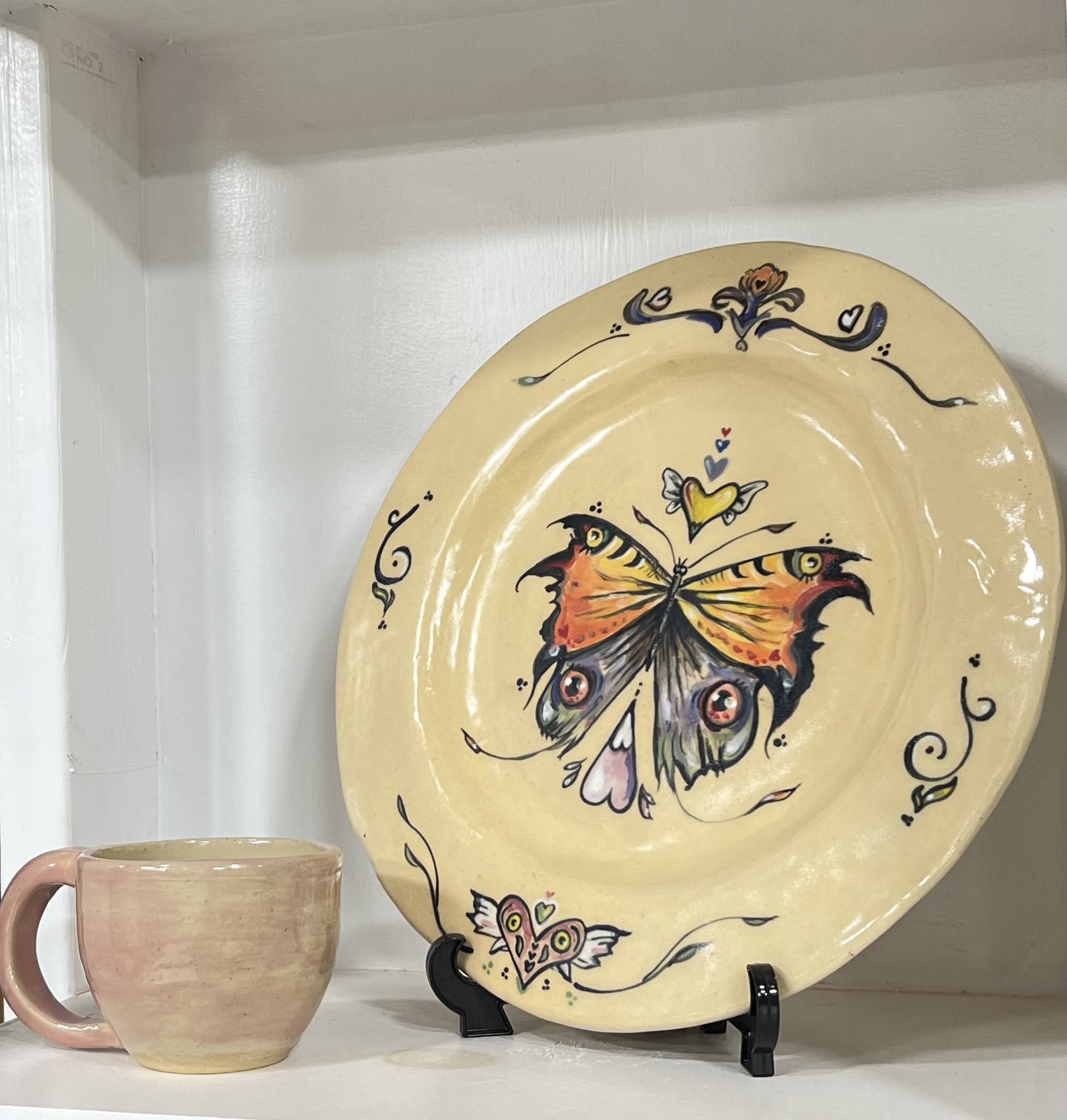 Decorative plate with a butterfly design, placed on a stand next to a ceramic mug, on a shelf.