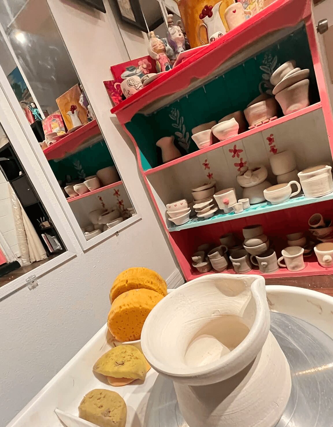 A white mortar and pestle on a tray with three yellow sponges or scrubbers beside it, in front of a pink and teal shelf filled with small bowls and cups, and a window with a reflection of a room.