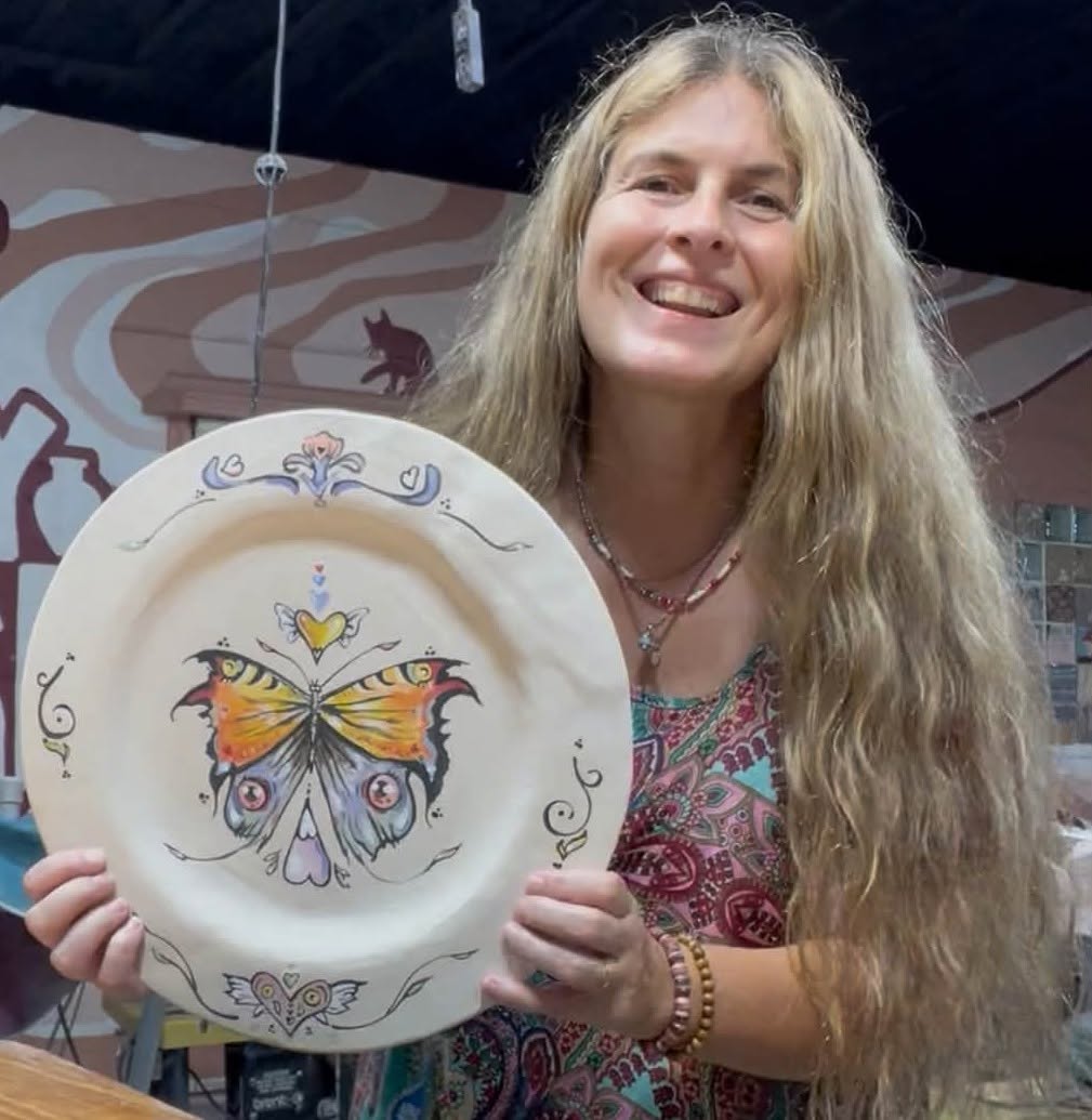 Woman with long hair smiling holding a decorative plate with a butterfly design, background with mural and mosaic tiles.