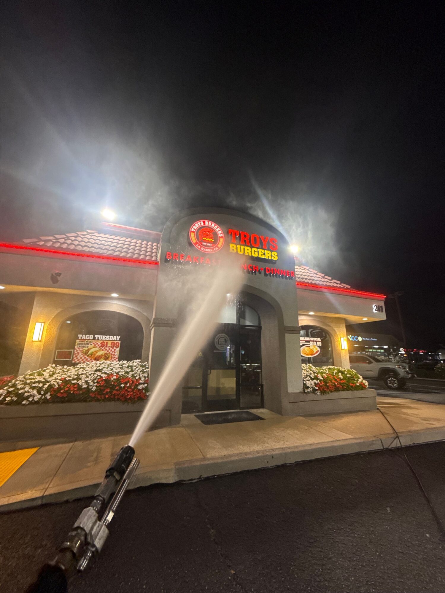 Nighttime scene of a gas station with a spray of water or foam, illuminated by bright lights, and flowers near the entrance.