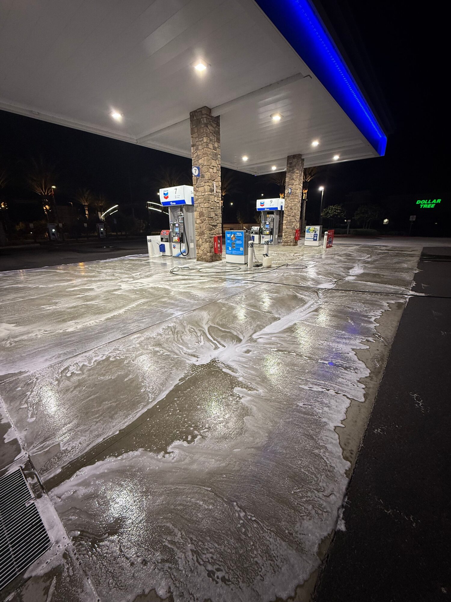 Street with wet pavement and illuminated gas station at night, with visible signs and structures.