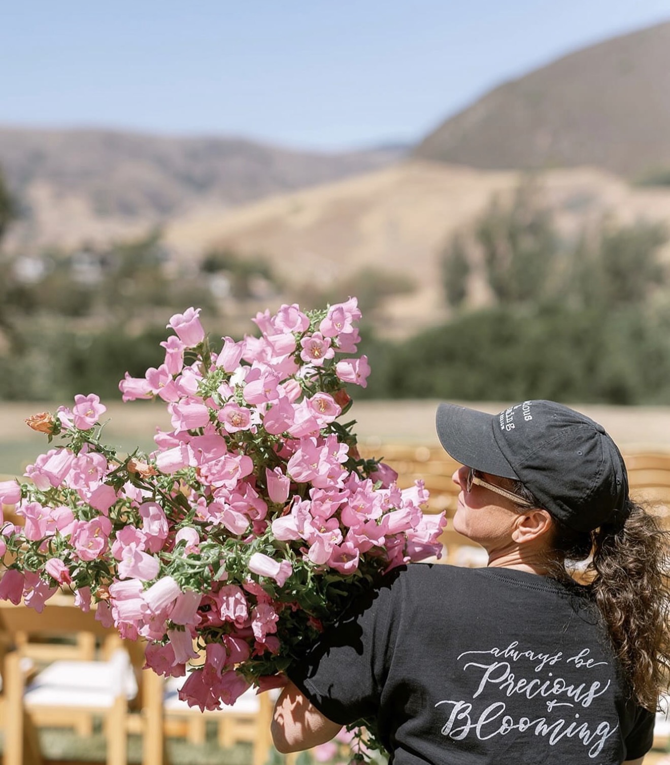 Person holding a large bouquet of pink flowers outdoors with hills and trees in the background, wearing a black cap and shirt.