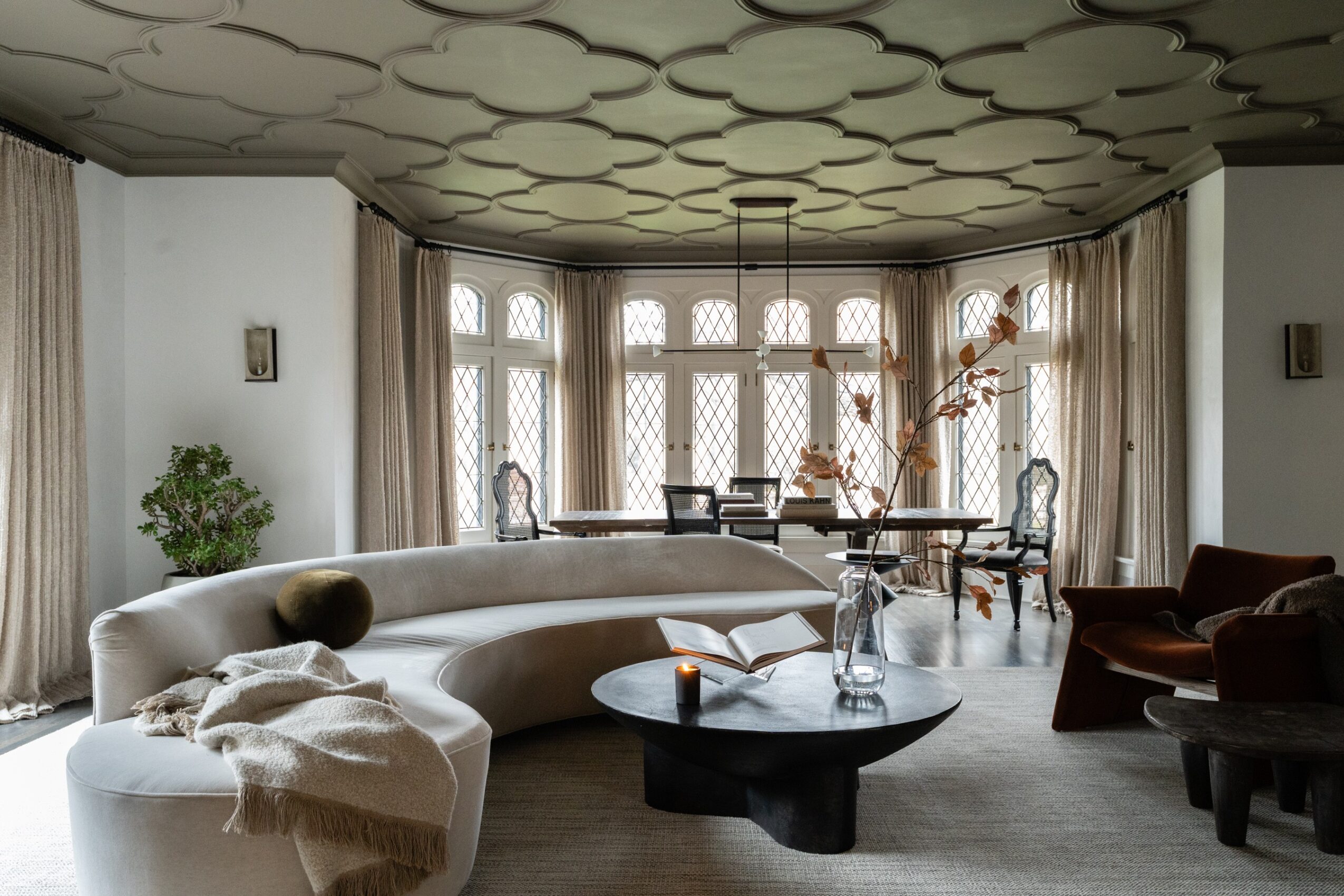 Living room with curved sofa, large windows, and decorative ceiling, featuring a coffee table and chairs, with curtains and plants.