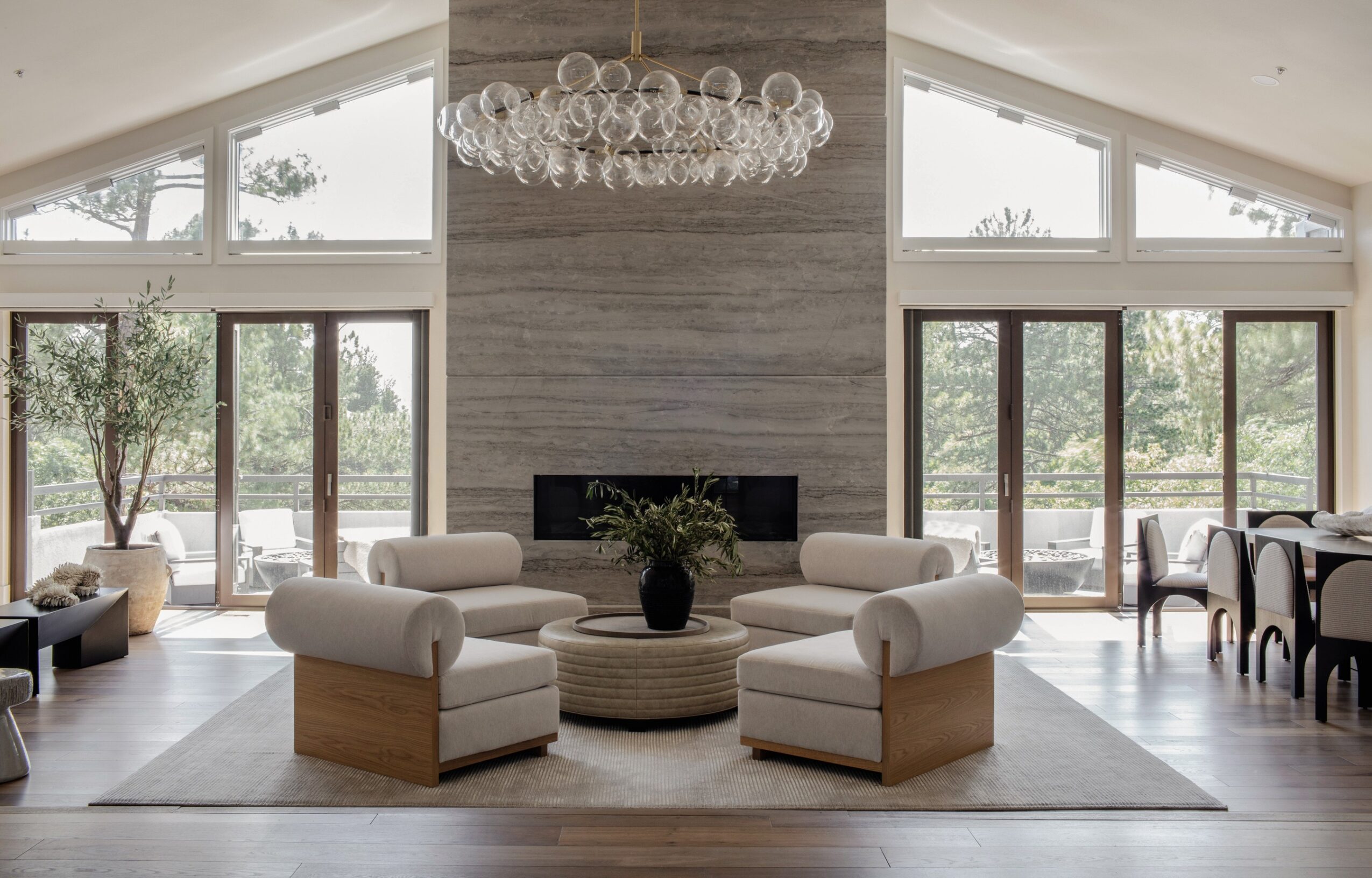 Living room with large windows, white sofas, a chandelier, and a fireplace, with outdoor view and dining area visible.