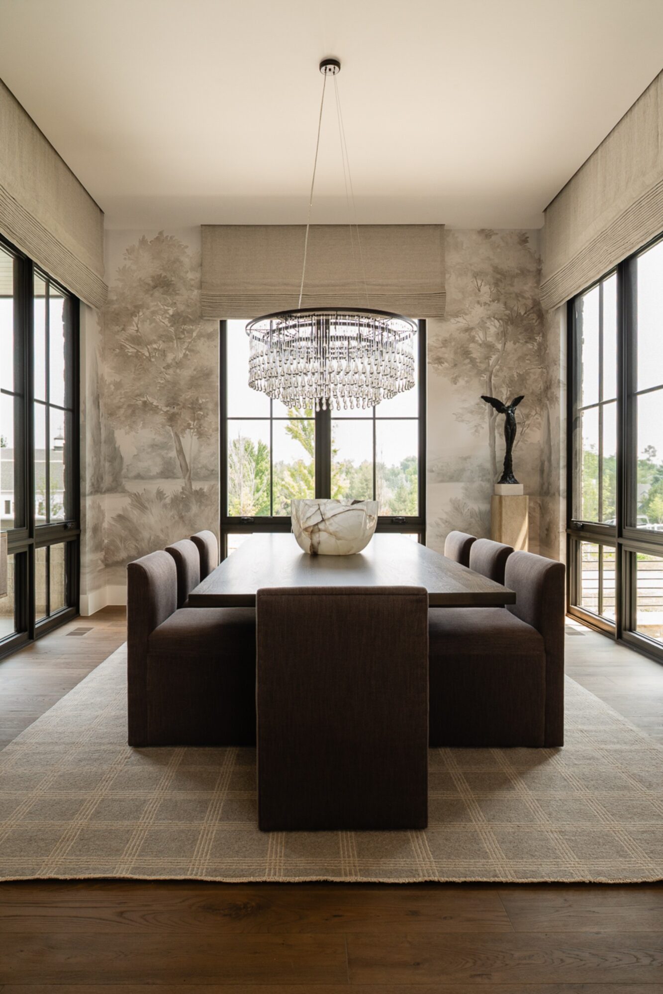 Dining room with large windows, chandelier, table, and chairs, decorated with a vase and sculpture, natural light, neutral tones.