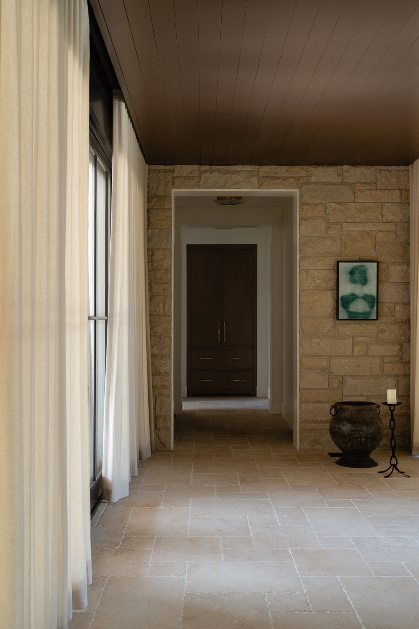 Interior hallway with stone wall, doorway, artwork, and decorative items, illuminated by natural light from large windows with curtains.