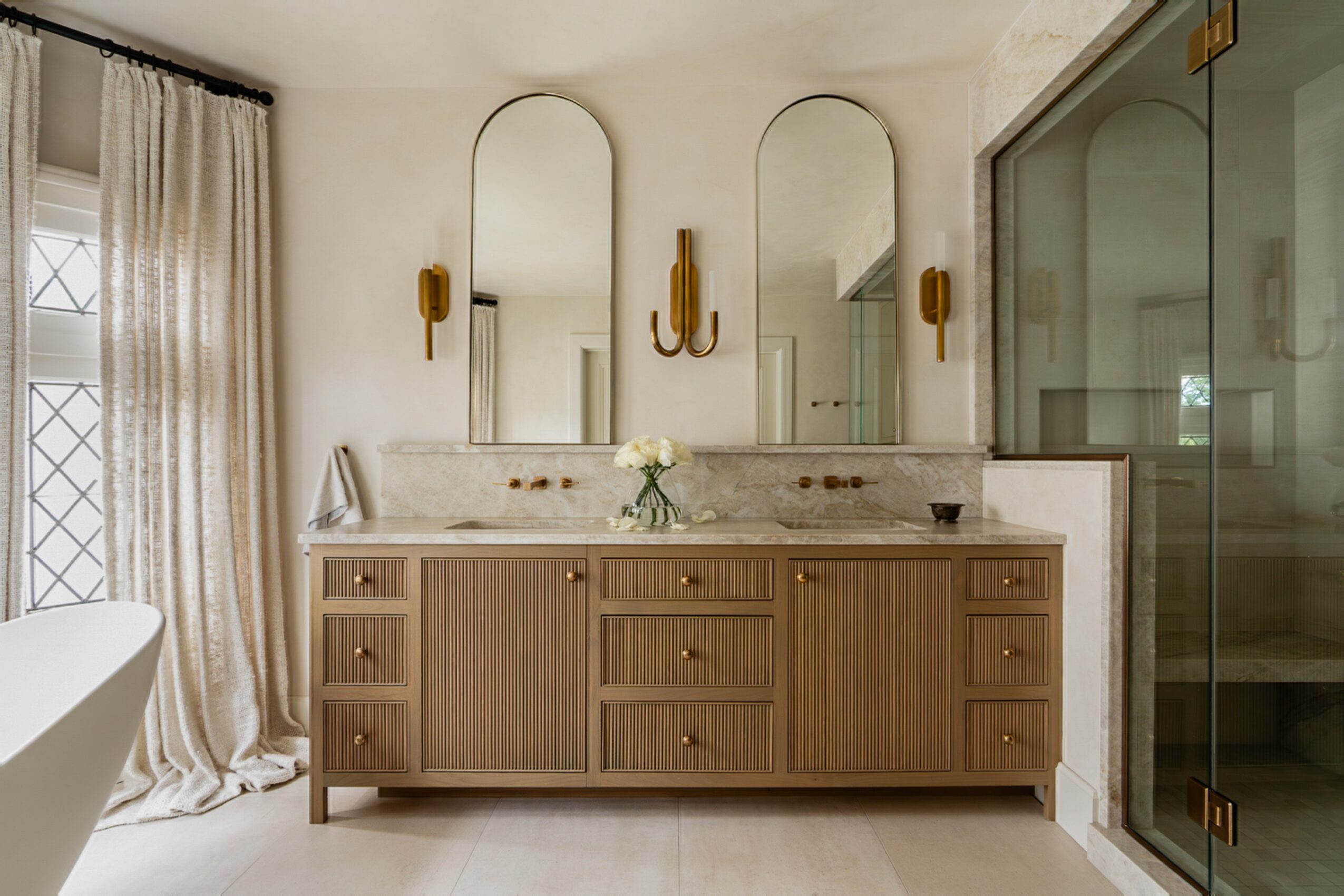 Bathroom vanity with two mirrors, wooden cabinet, flower vase, and a shower enclosure on the right.