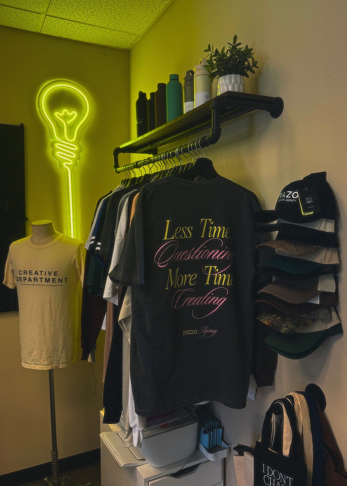 Clothing store interior with a neon light of a lightbulb on the wall, shelves with bottles, and a rack of shirts.