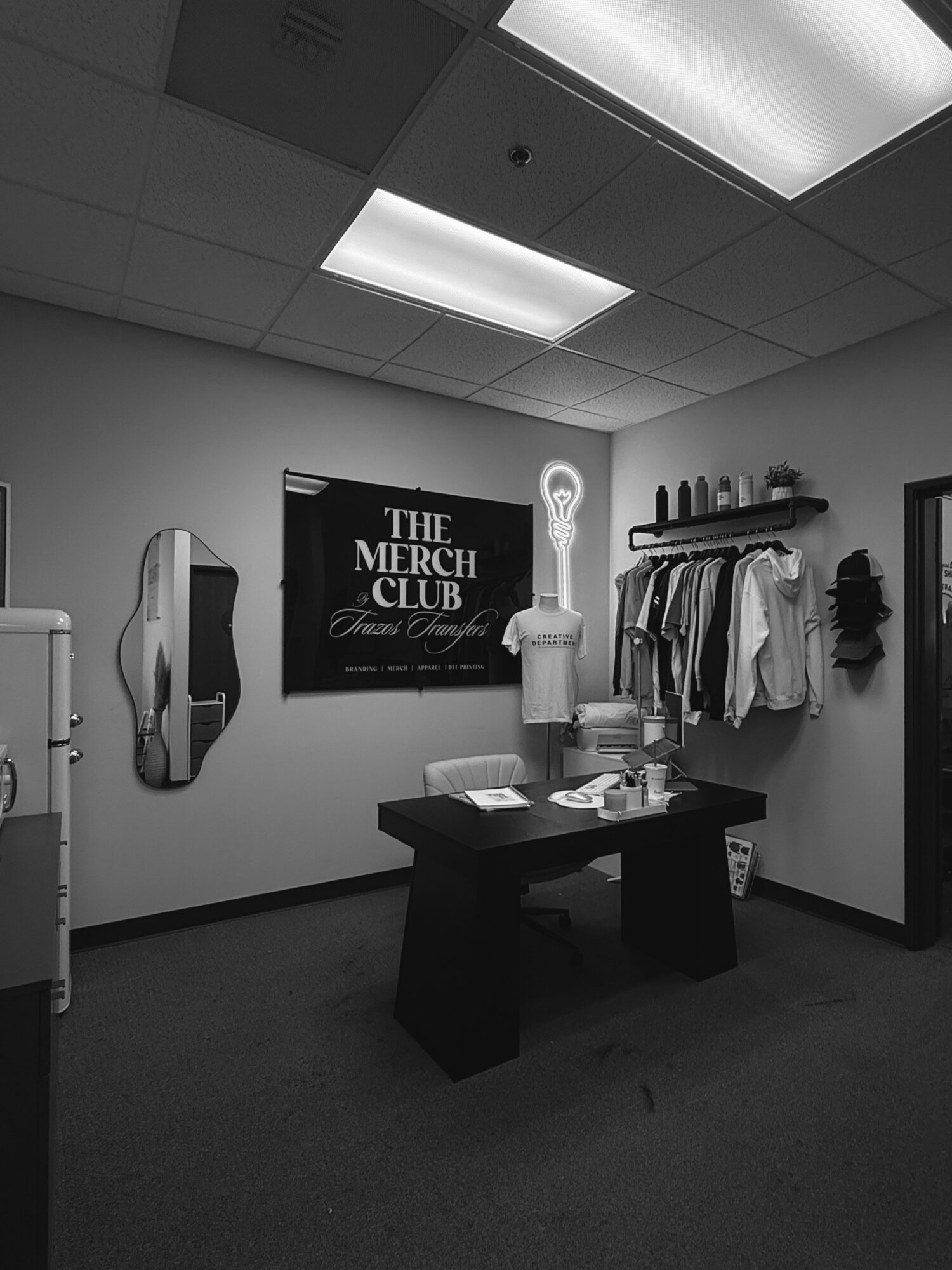 Clothing display with shirts, a table with papers, and a sign reading 'The Merch Club' in a room with ceiling lights.