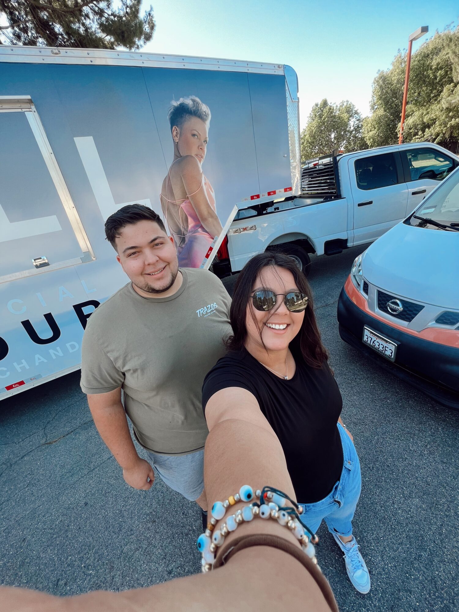 Two people smiling in a parking lot, with a truck and a car behind them, and a large advertisement on the truck.