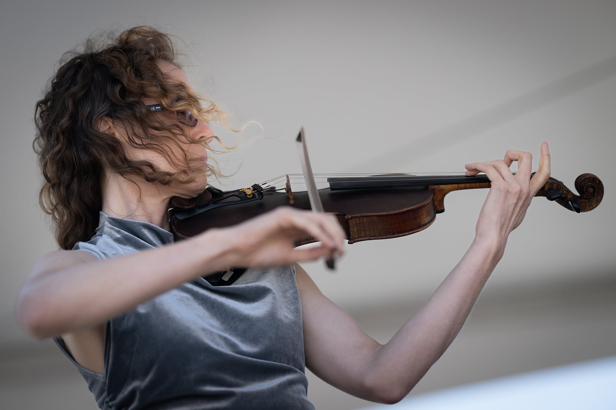 Person with curly hair playing a violin, wearing a shiny sleeveless top, with a neutral background.