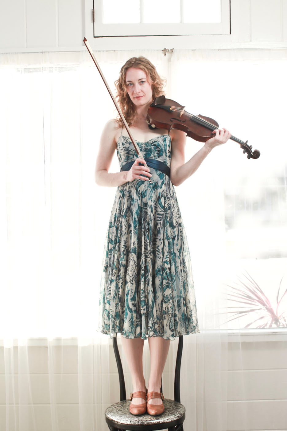 Woman standing on a stool playing a violin indoors, wearing a patterned dress and brown shoes.