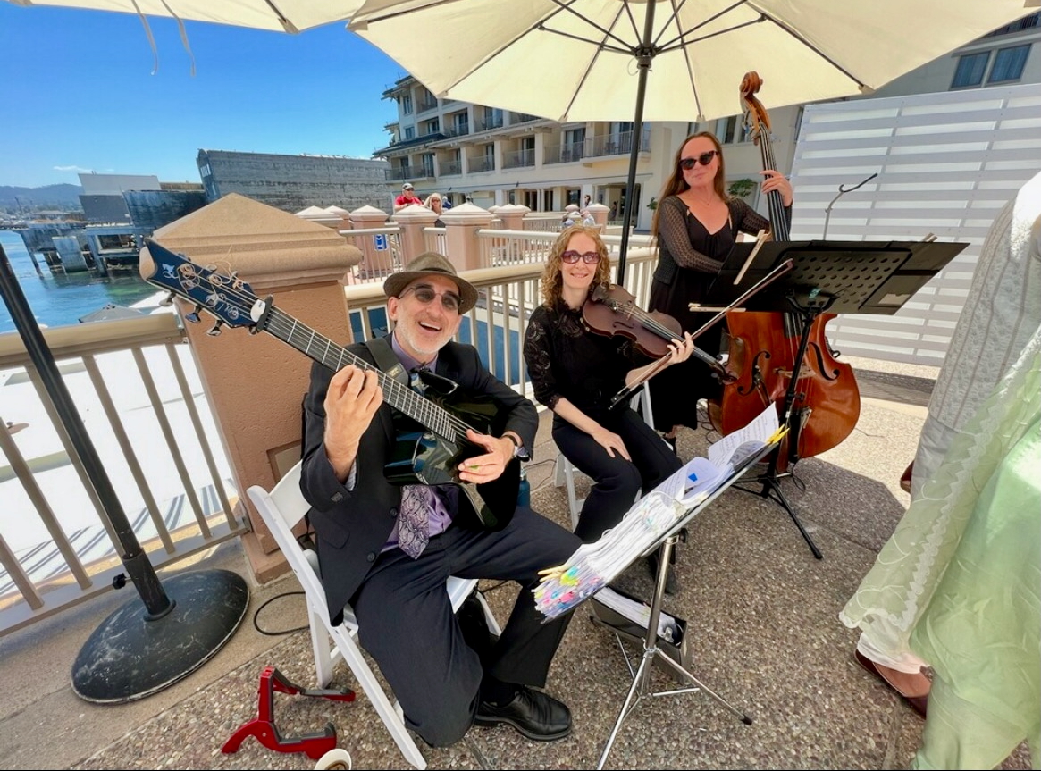 Three musicians playing instruments outdoors under a large umbrella, with a waterfront and buildings in the background.