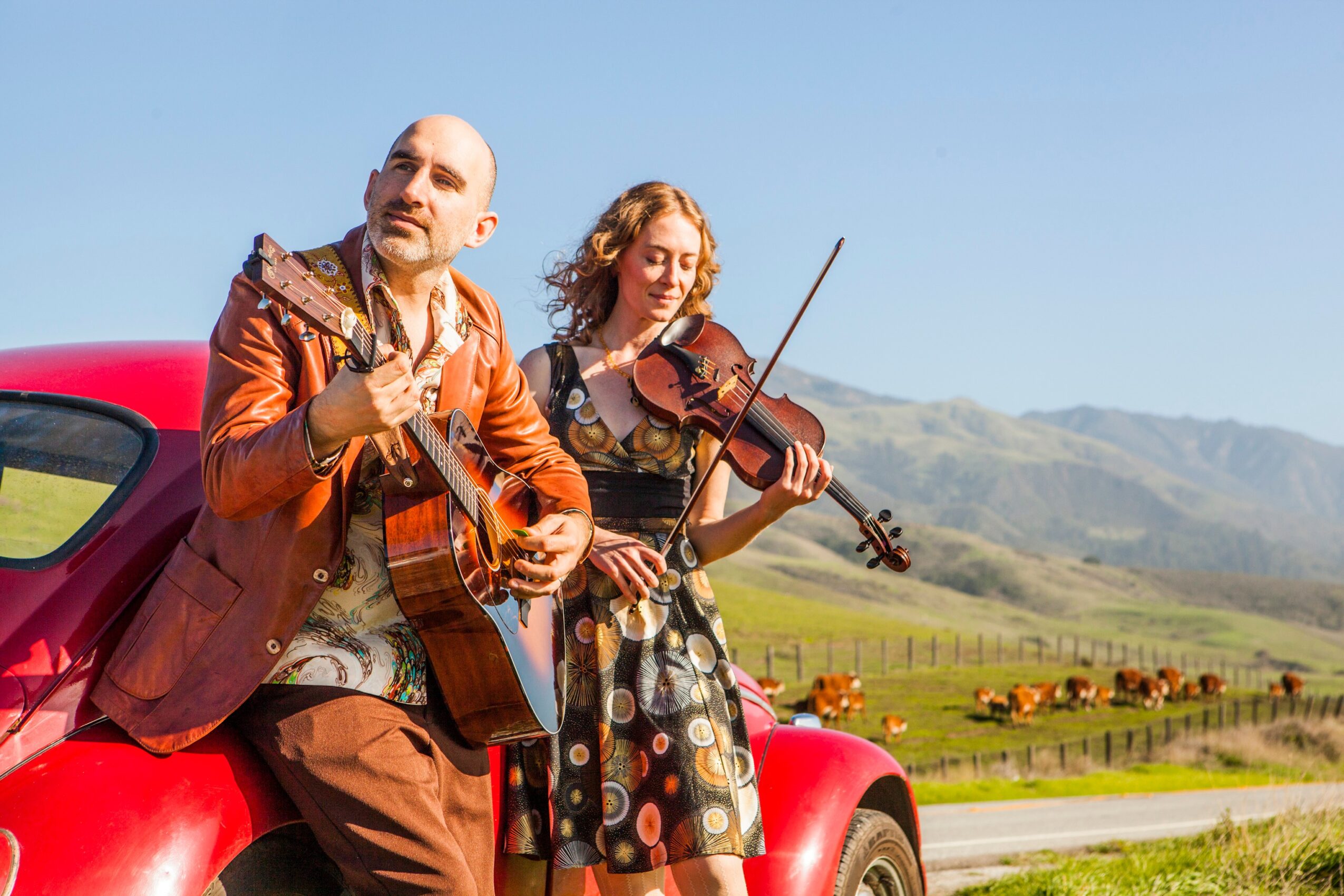 Two musicians, a man with a guitar and a woman with a violin, sit on a red car outdoors with hills in the background.