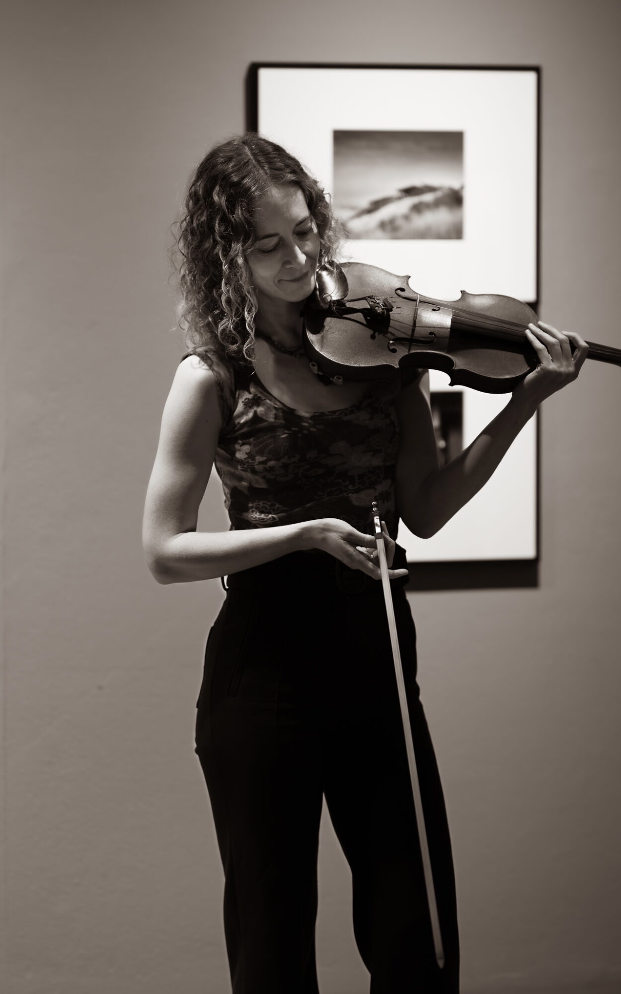 Woman playing violin indoors, standing in front of framed artwork, with curly hair and glasses, wearing a sleeveless top and pants.