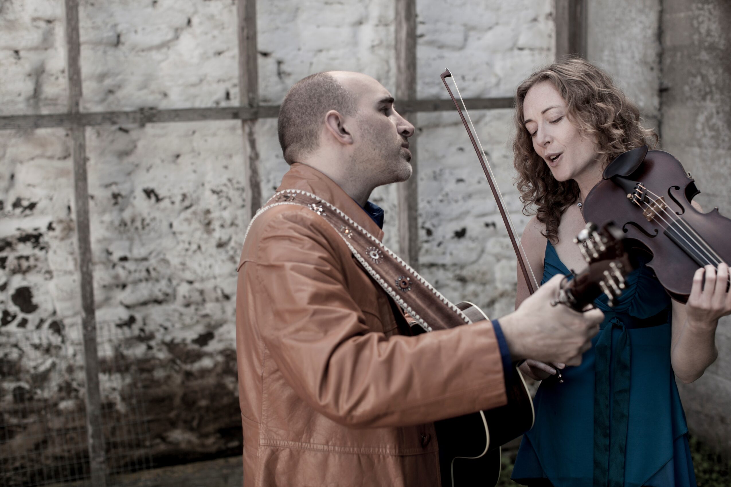 Two people playing musical instruments outdoors against a stone wall, one with a guitar and the other with a violin, engaging in a duet.