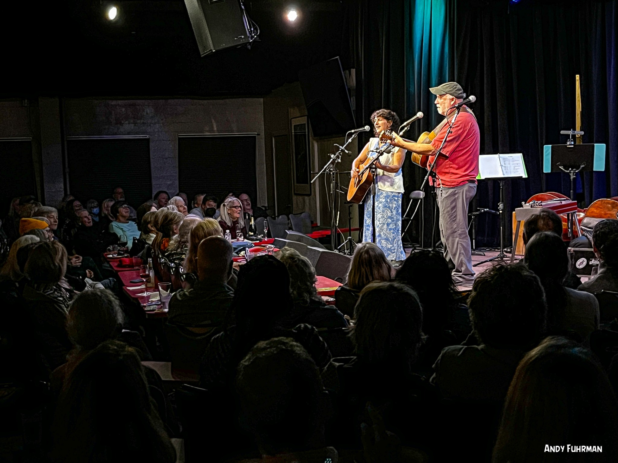 Two musicians perform on stage with an audience watching, one playing guitar and the other singing, in a dimly lit venue.