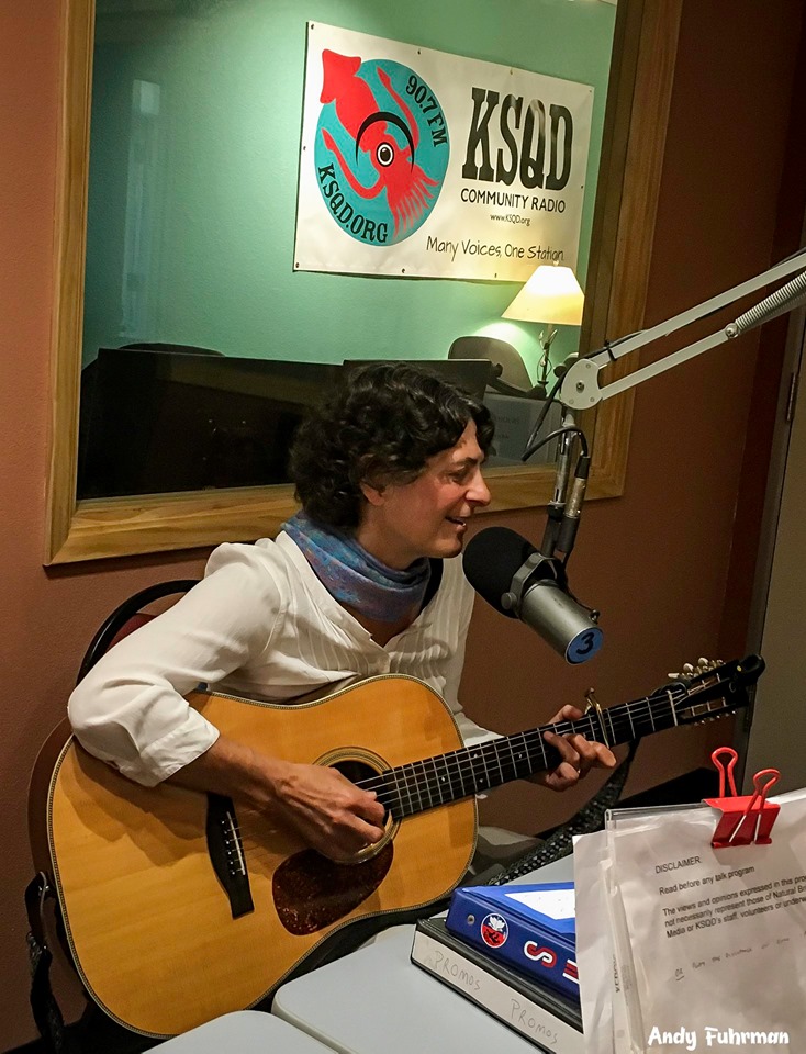 Person with curly hair playing guitar and singing into a microphone in a radio studio, with a KSKD Community Radio sign in the background.