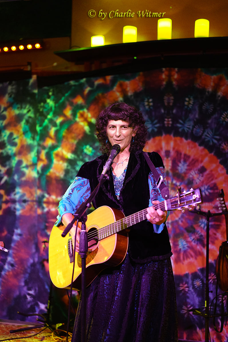 Person with curly hair playing guitar and singing on stage with colorful tie-dye backdrop.