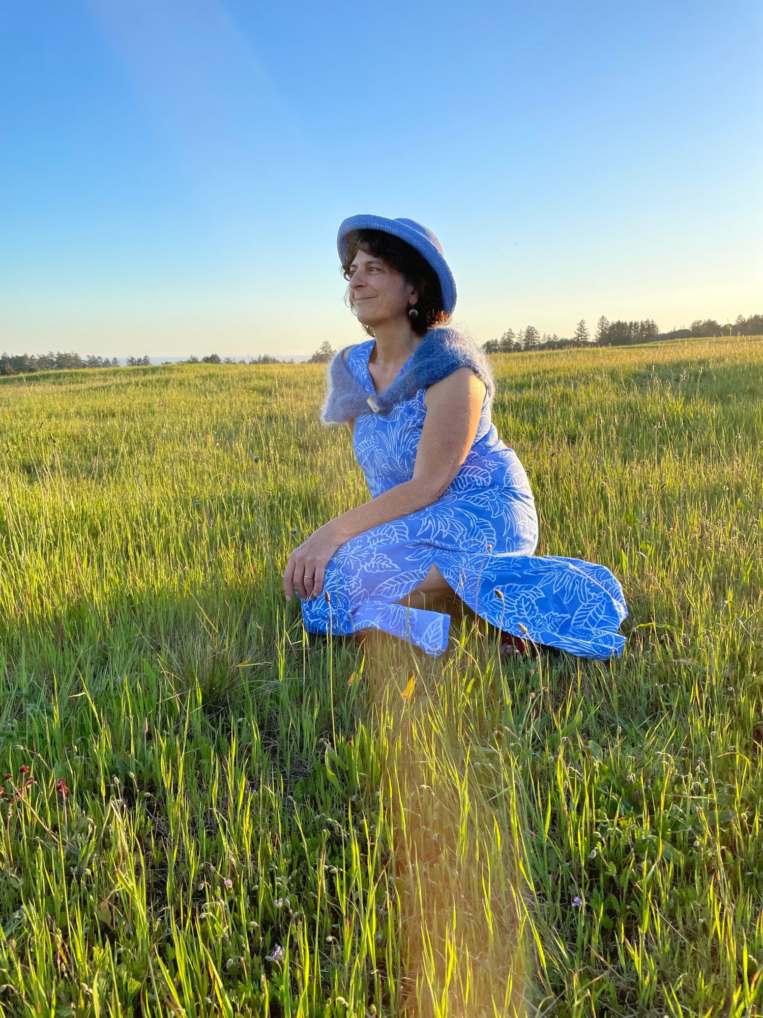 Woman in blue dress and hat kneeling in a green field under a clear blue sky.