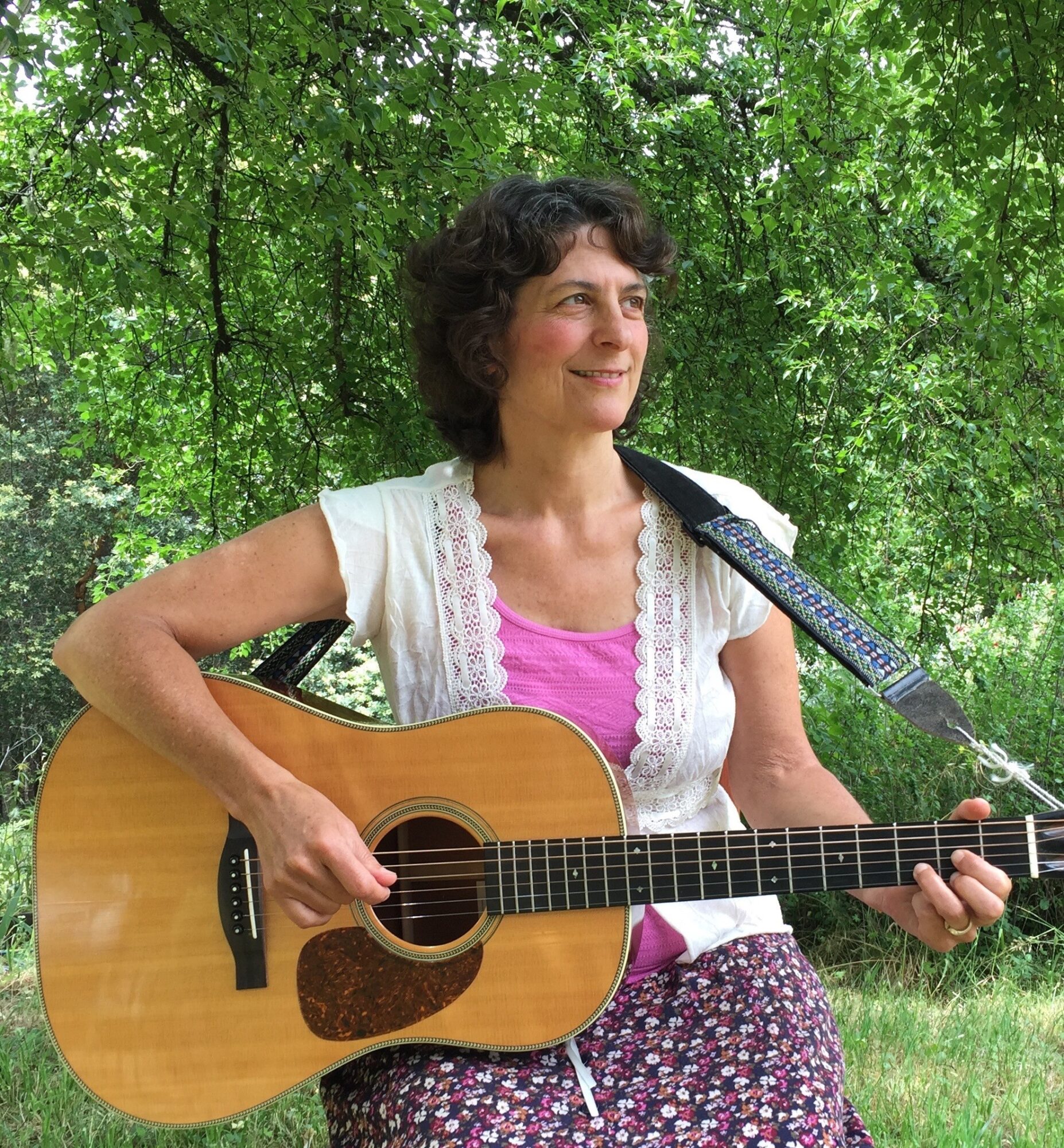Woman playing guitar outdoors with green trees in background, smiling, wearing a pink top and white vest.