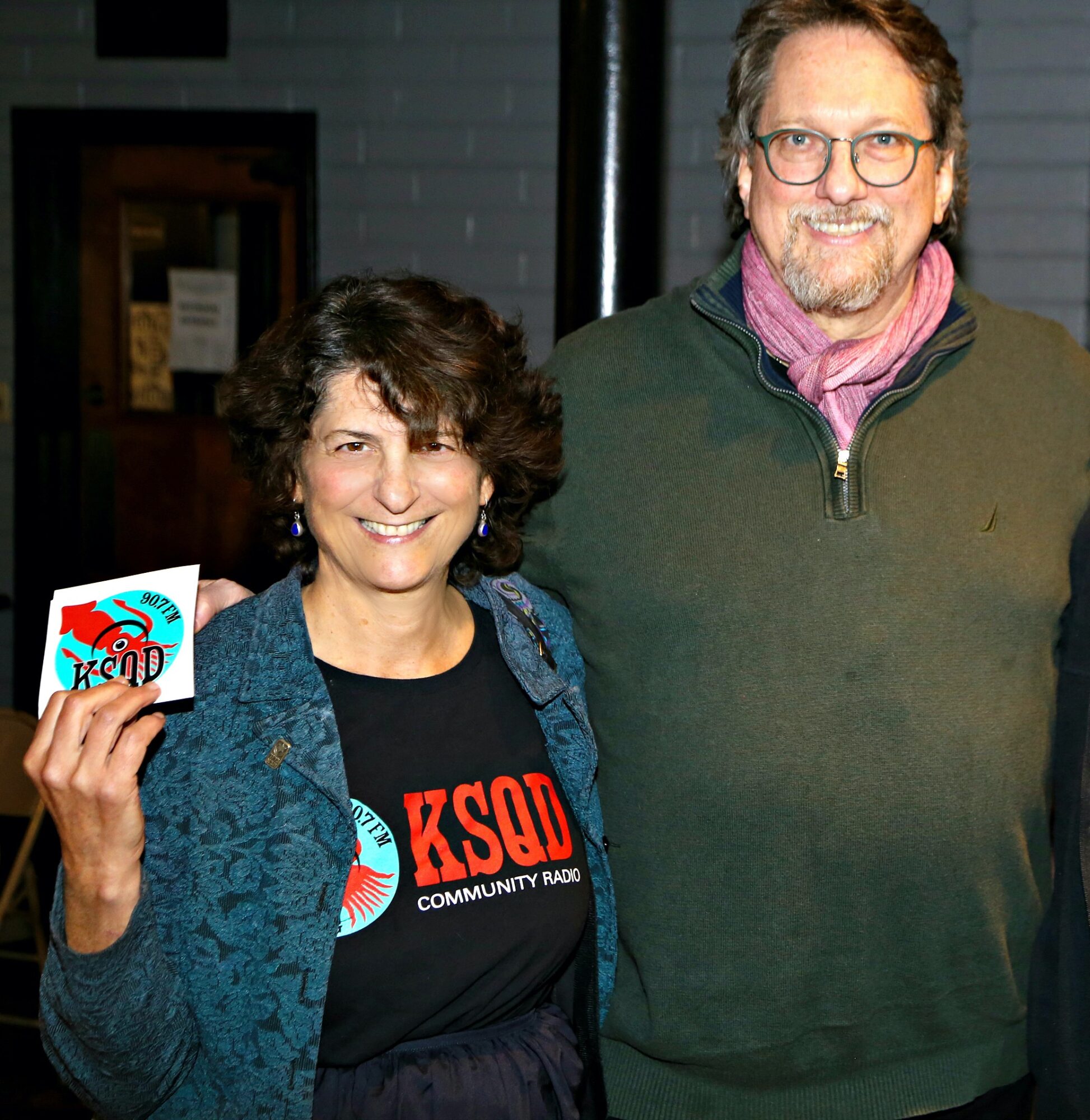 Two people smiling, woman holding a card, woman wearing a black shirt with red text, man in green jacket with pink scarf