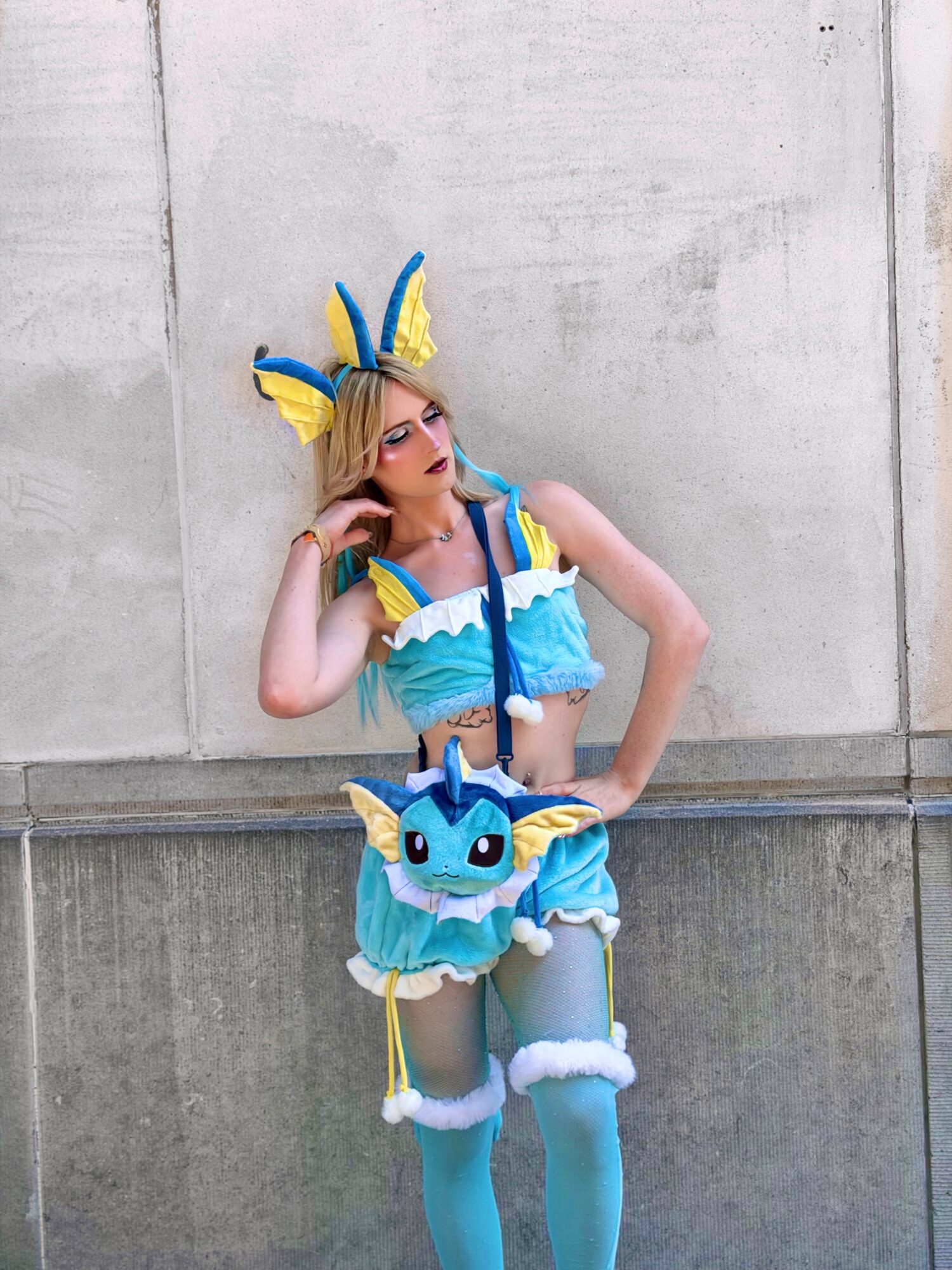 Person in colorful costume with blue, yellow, and white elements, wearing a headpiece with large ears, standing against a concrete wall.
