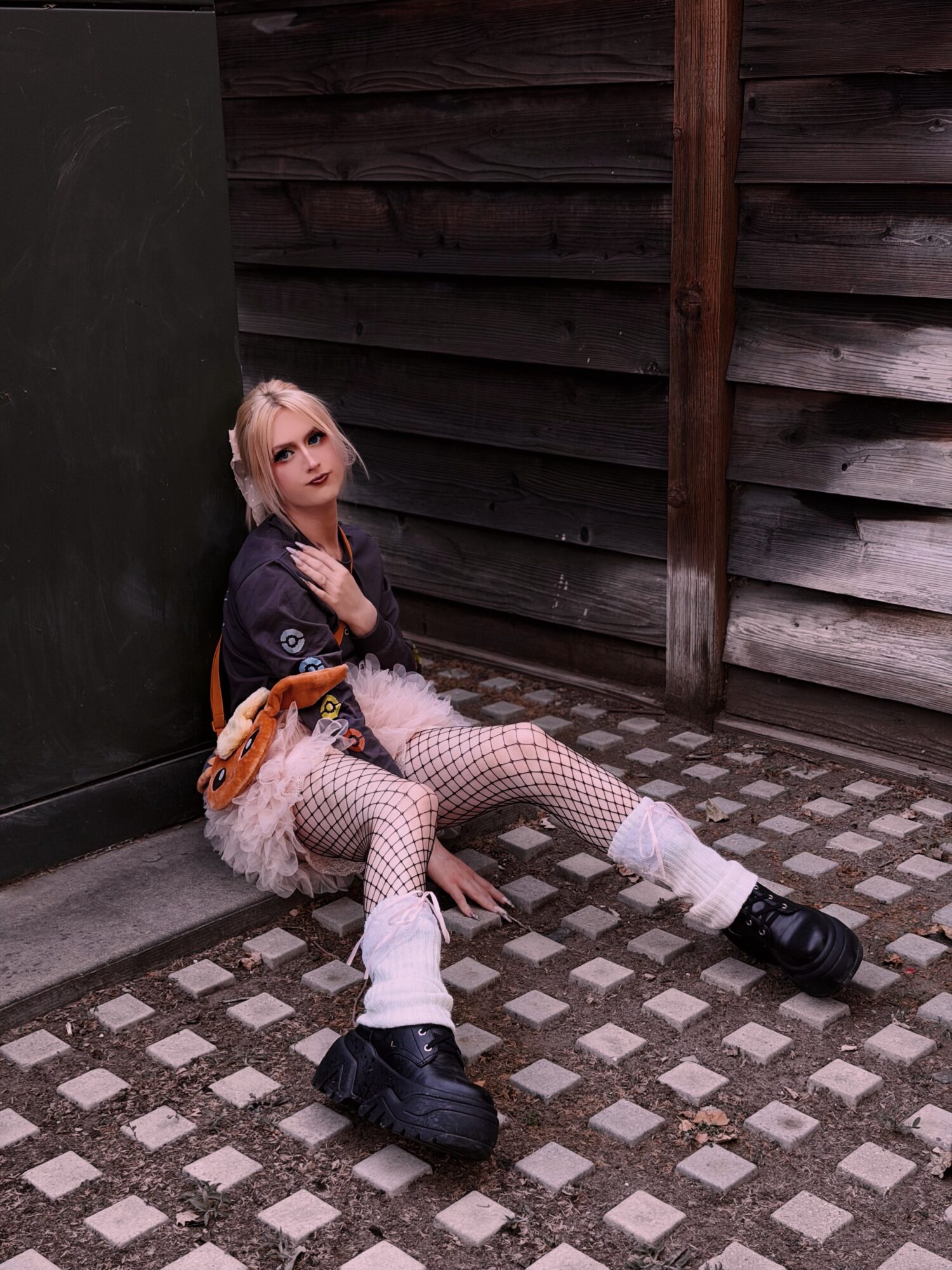 Person sitting on ground against a wall, looking up, with a wooden fence behind, wearing patterned pants and black shoes.