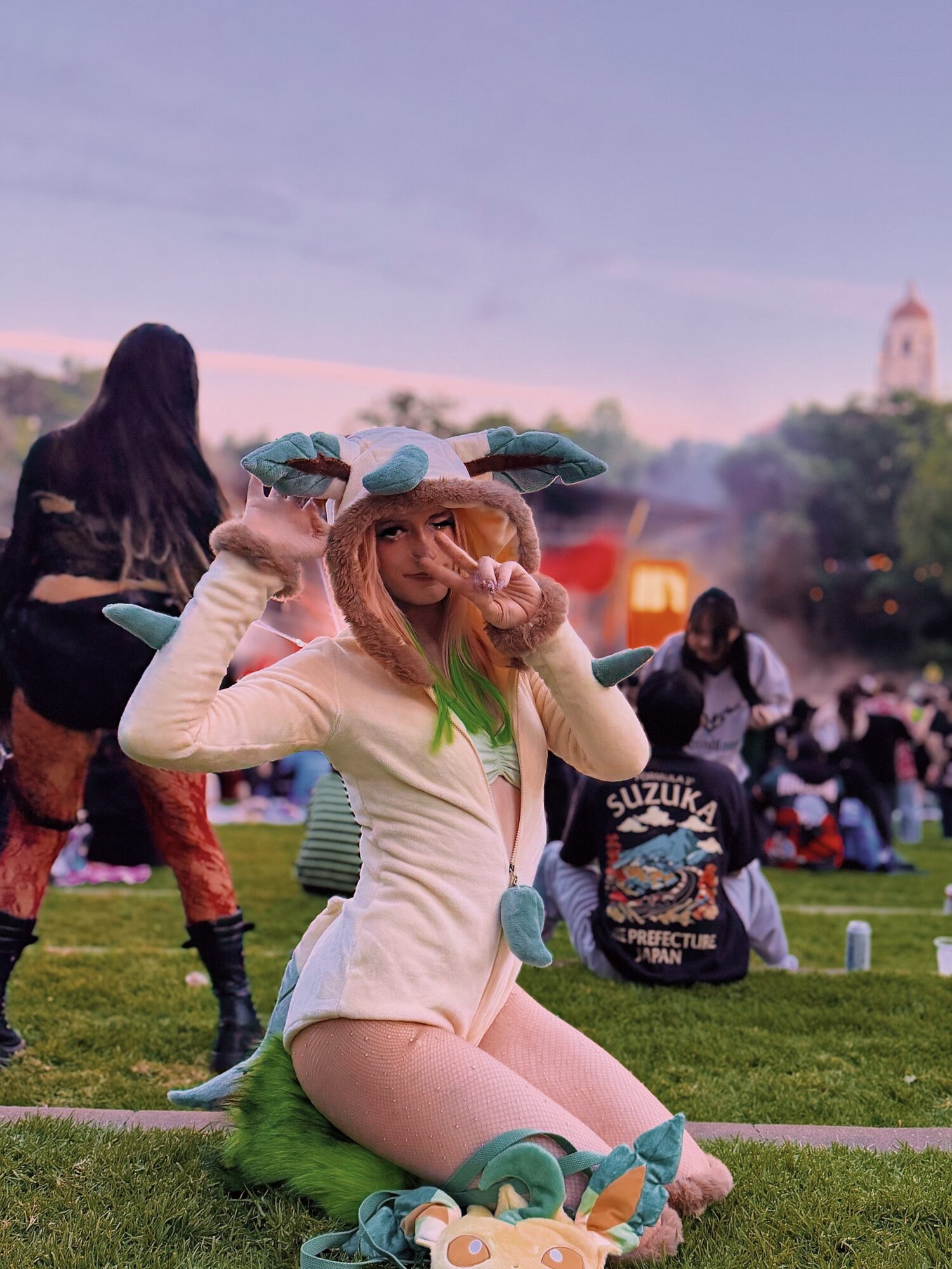 Person in a costume with animal ears and tail, sitting on grass at outdoor event, surrounded by people and trees.