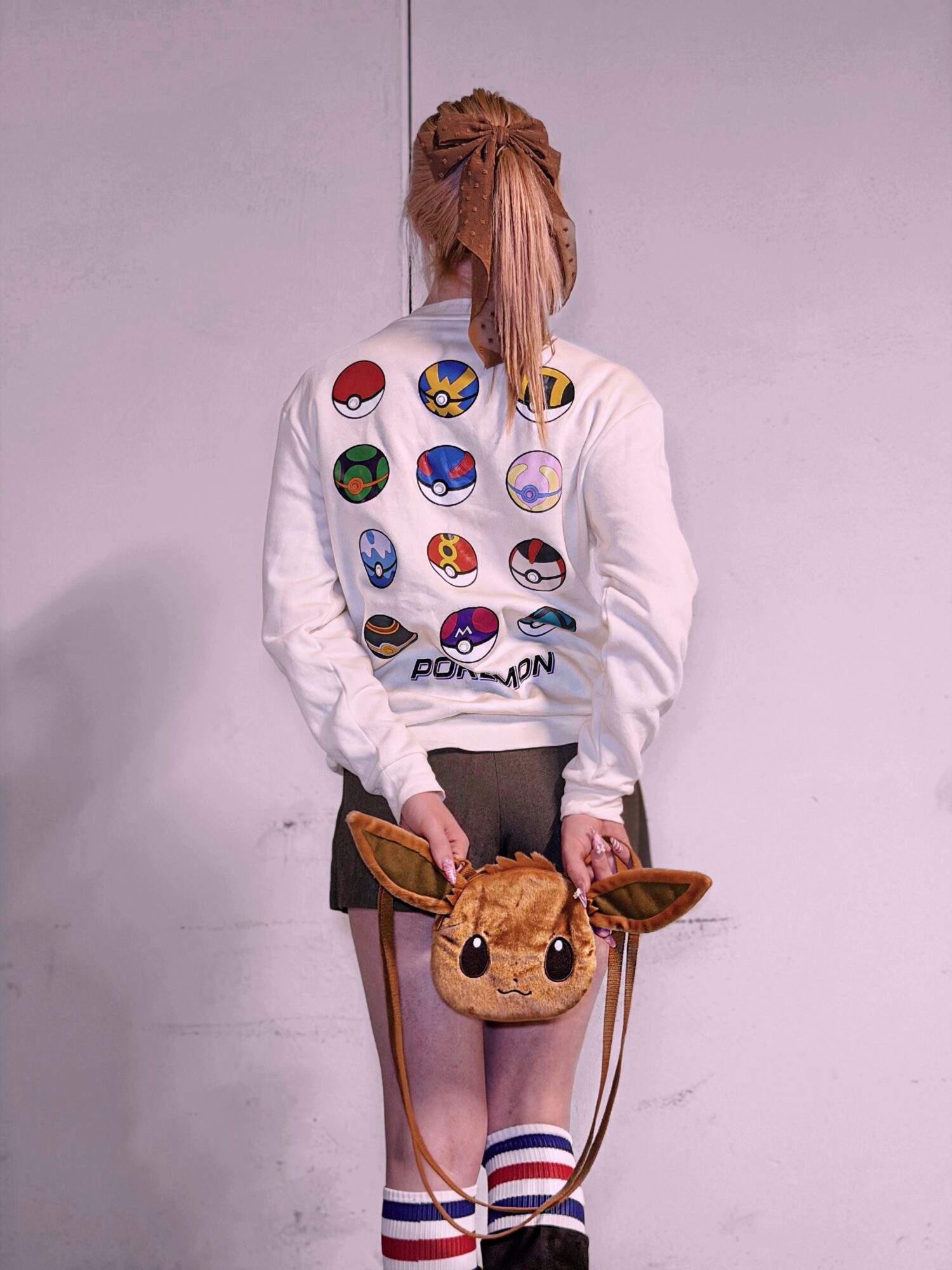 Person with long hair holding a plush Eevee bag, wearing a white sweatshirt with Pokémon logos, striped socks, and shorts.