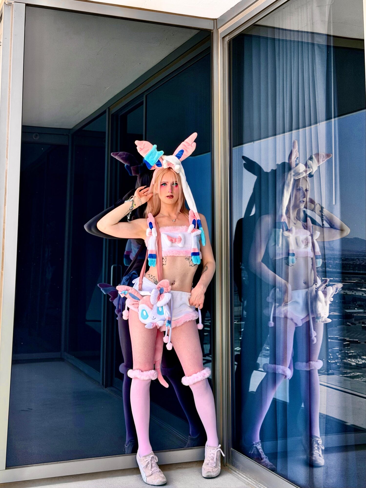Person dressed as a unicorn with pastel-colored accessories, standing indoors near a glass window with a reflection, ocean view outside.
