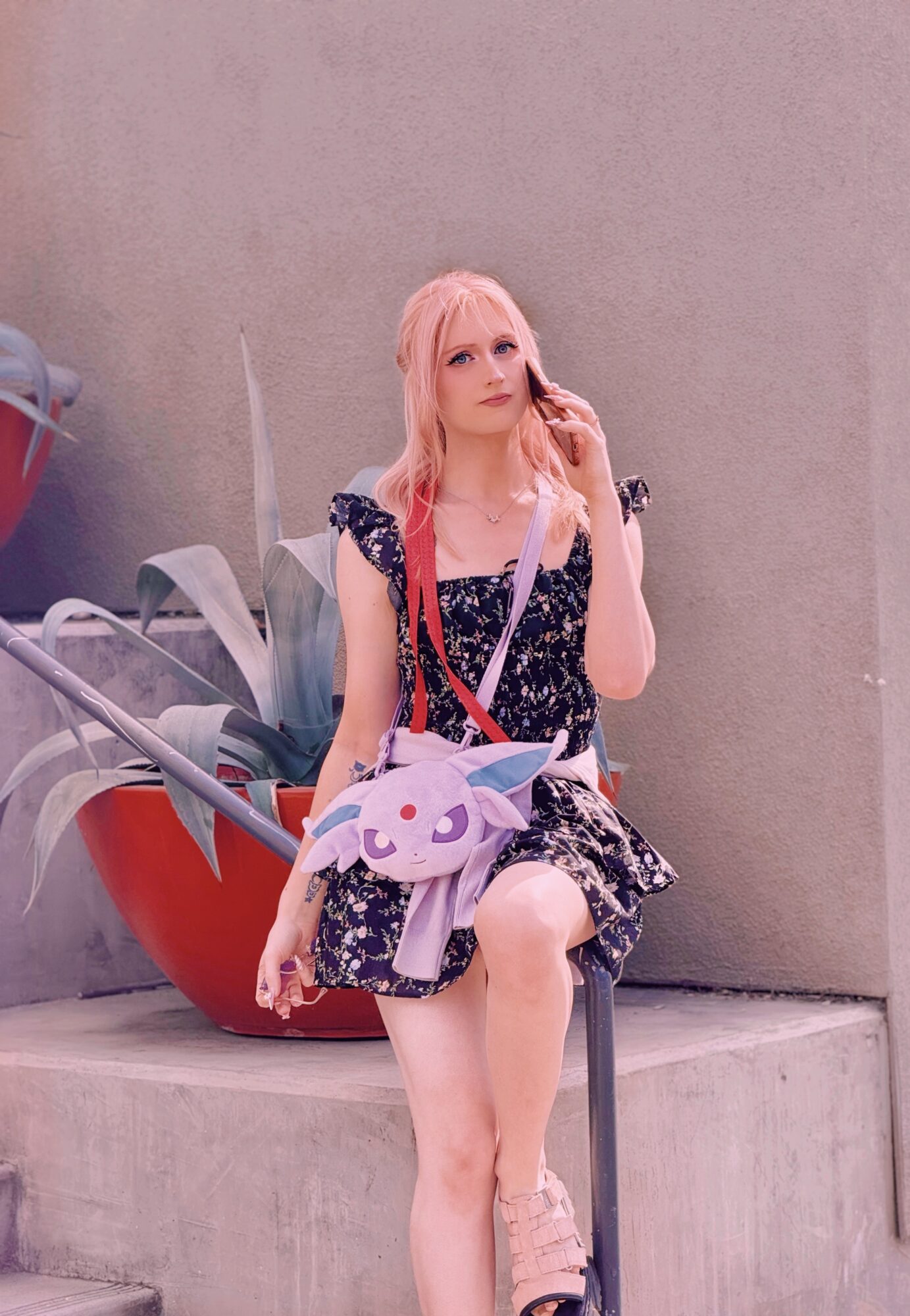 Young woman with pink hair sitting on a ledge, holding a plush unicorn bag, against a plain wall.