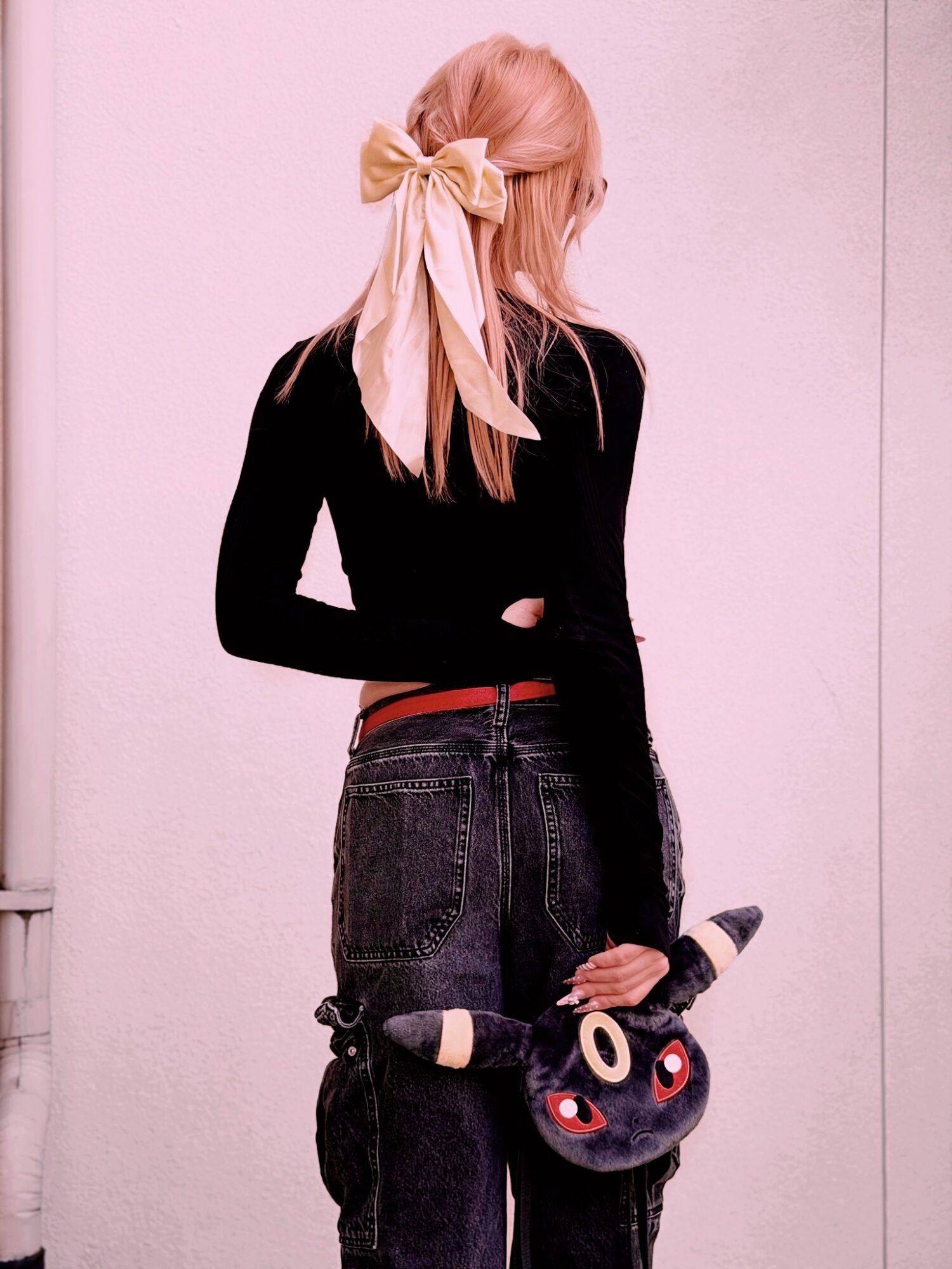Person with long blonde hair tied with a large yellow bow, wearing a black top and dark jeans, holding a plush toy.
