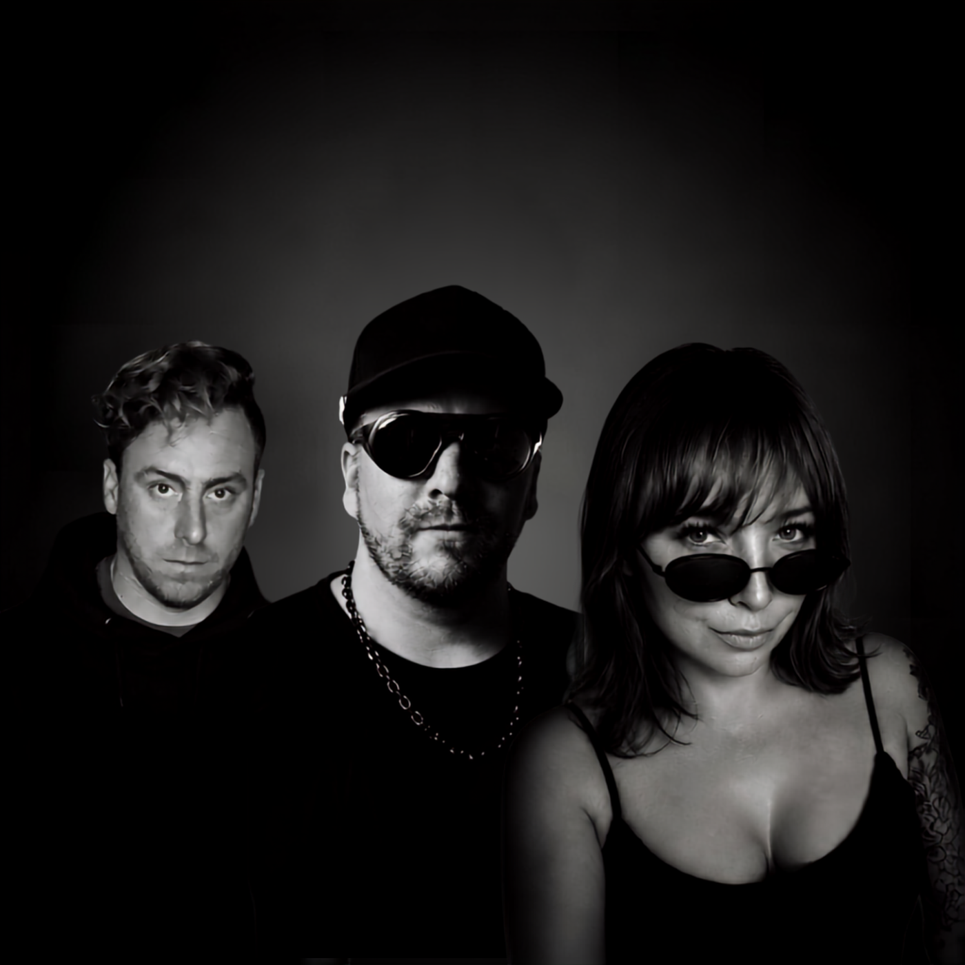Three people pose together, wearing dark clothing and sunglasses, against a dark background.