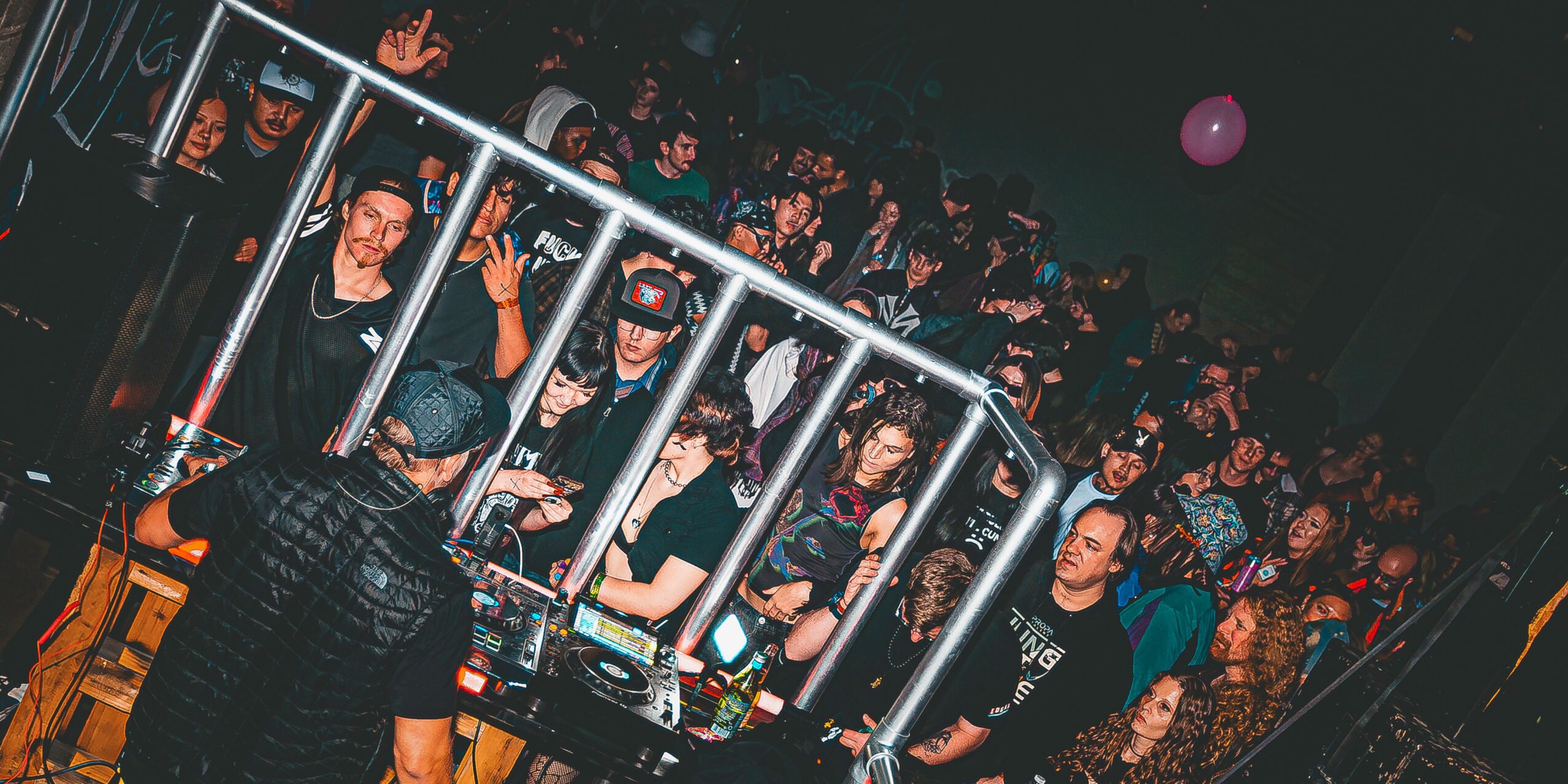 Crowd of people at a party or concert, some holding drinks, with a metal railing in the foreground, dim lighting.