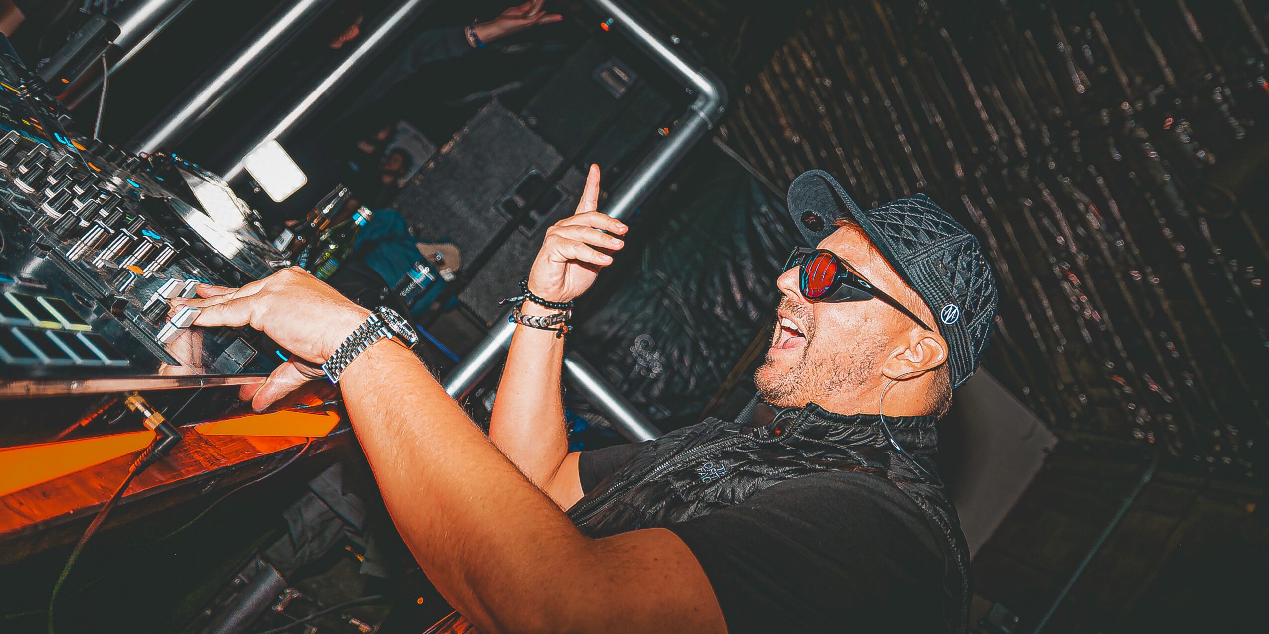 Person wearing sunglasses and a cap, smiling and pointing upward, standing near DJ equipment in a dark setting.