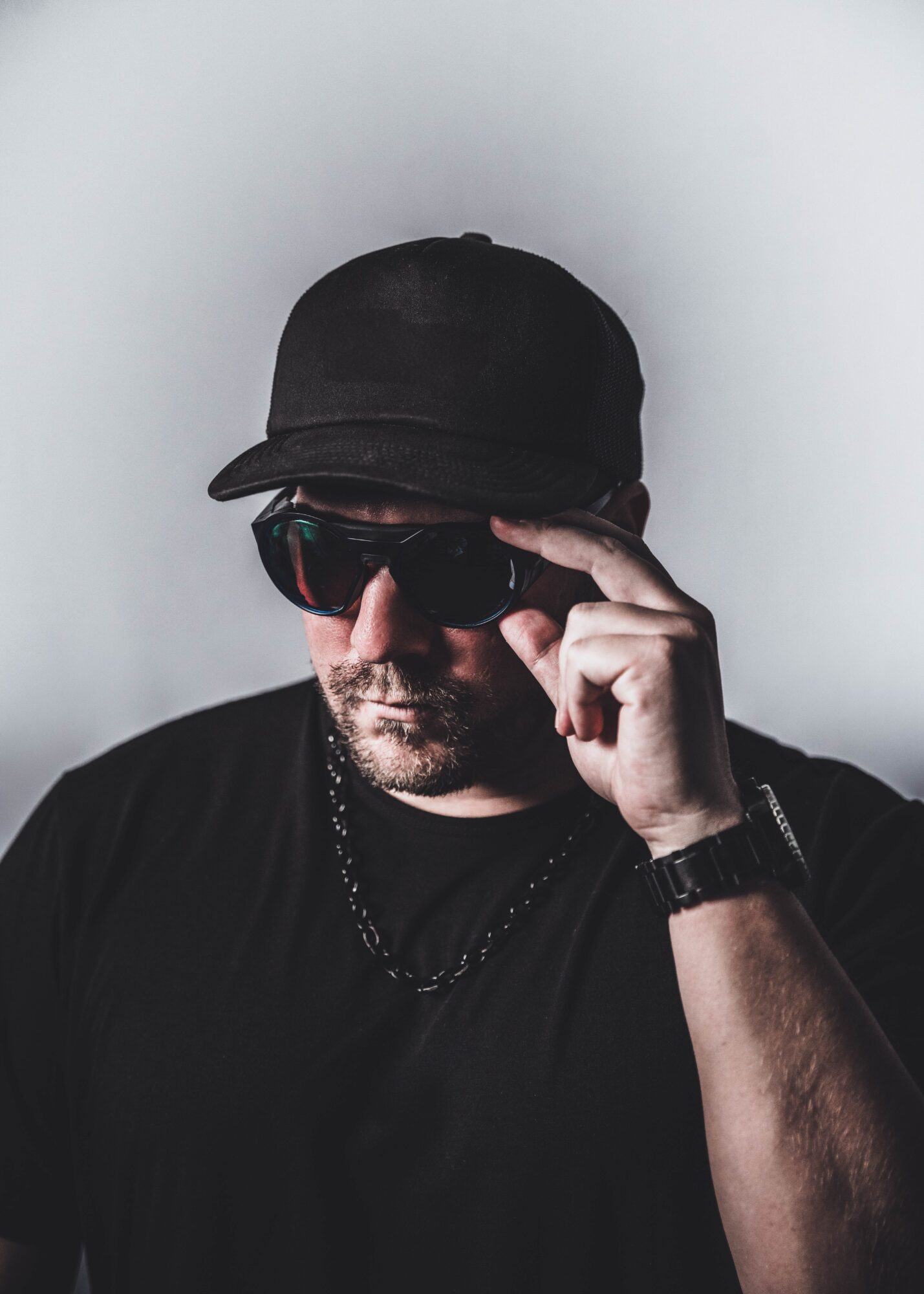 Man wearing a black cap and sunglasses, adjusting his sunglasses with his right hand, against a plain background.