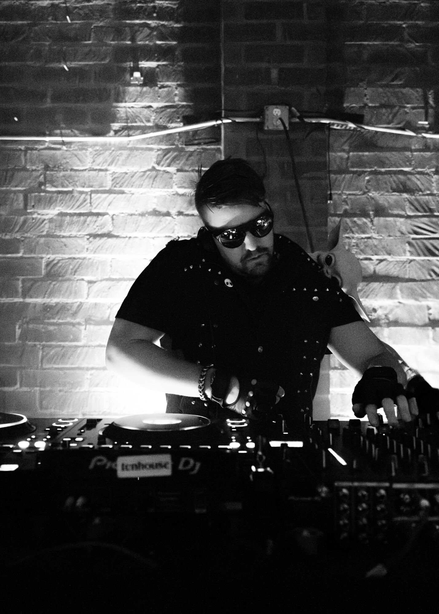 DJ wearing sunglasses and headphones, operating equipment against a brick wall, illuminated by a light source, in black and white.
